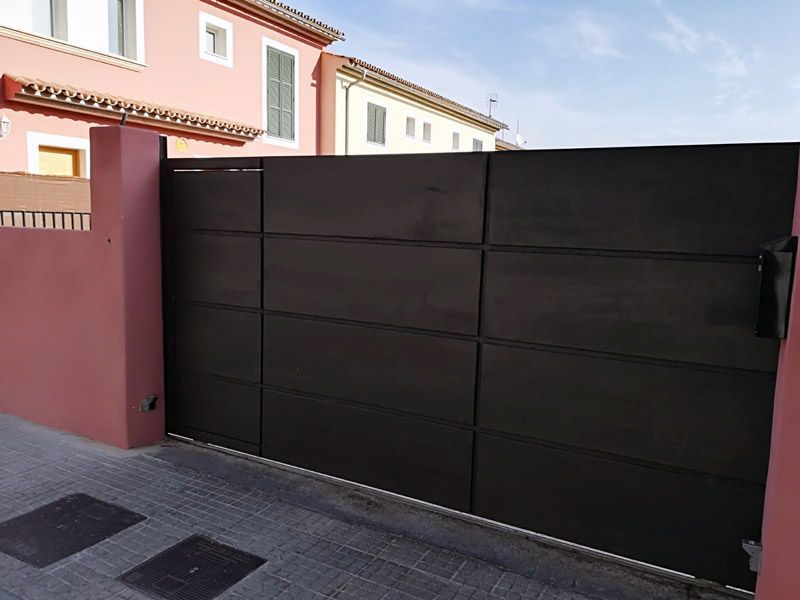 Puerta de metal negra frente a un edificio rosa y casas residenciales, protegiendo un camino de entrada.