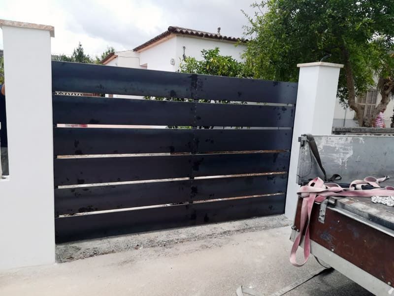 Puerta de lamas horizontales de color negro entre dos columnas blancas, camino de entrada de hormigón.