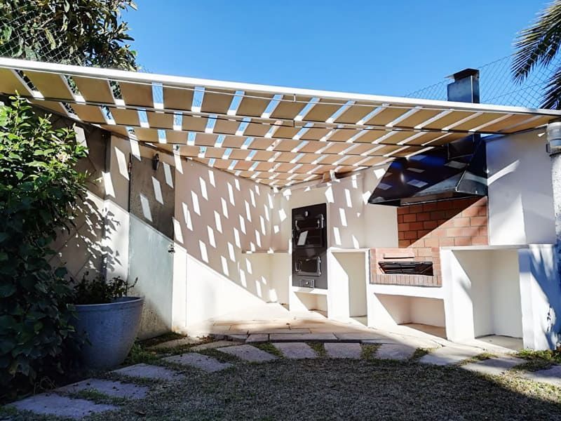 Patio exterior con toldo beige, parrilla empotrada y sendero de piedra. Día soleado.