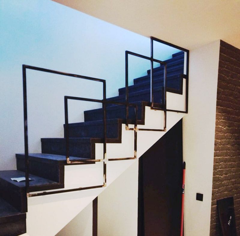 Escalera con barandilla metálica negra y geométrica. Peldaños de moqueta azul oscuro y paredes blancas.