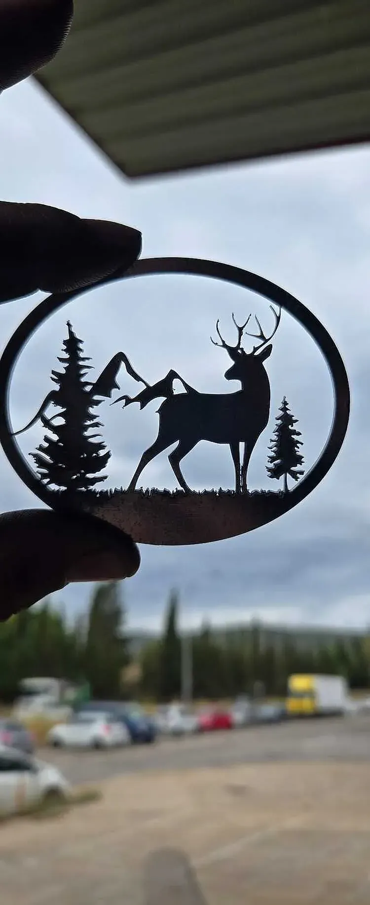 Silueta de un ciervo en un marco circular con montañas y árboles, sostenida frente a una escena borrosa al aire libre.