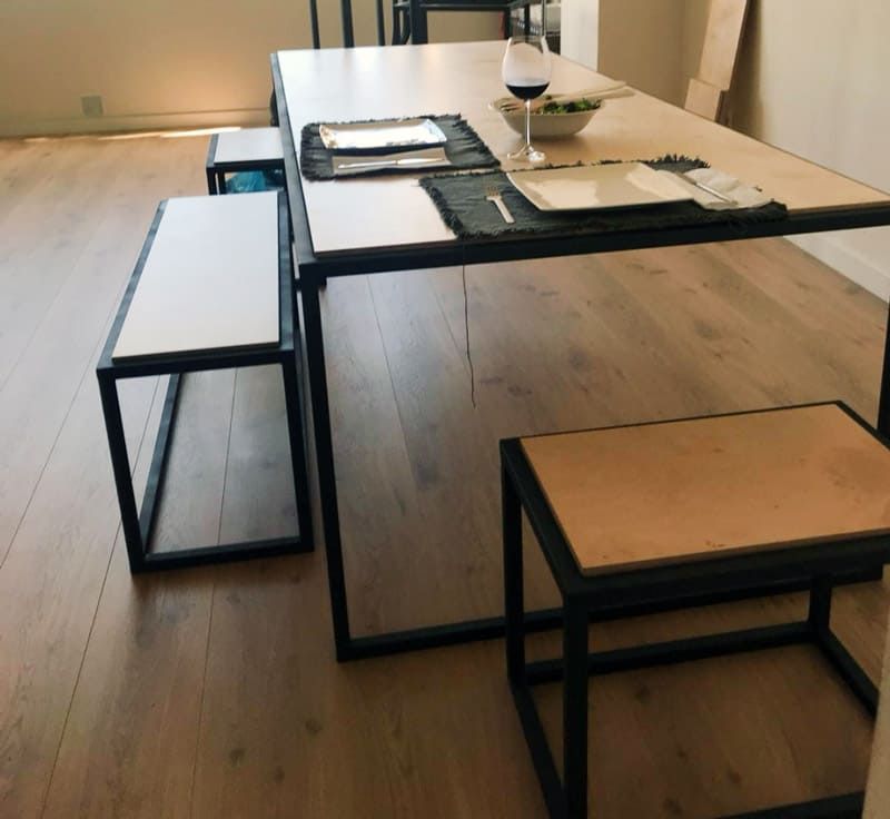 Mesa de comedor con estructura de metal y dos bancos, uno de ellos un taburete. Suelo de madera clara.