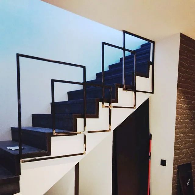 Barandilla de metal geométrica negra junto a escaleras alfombradas de color gris en una habitación blanca.