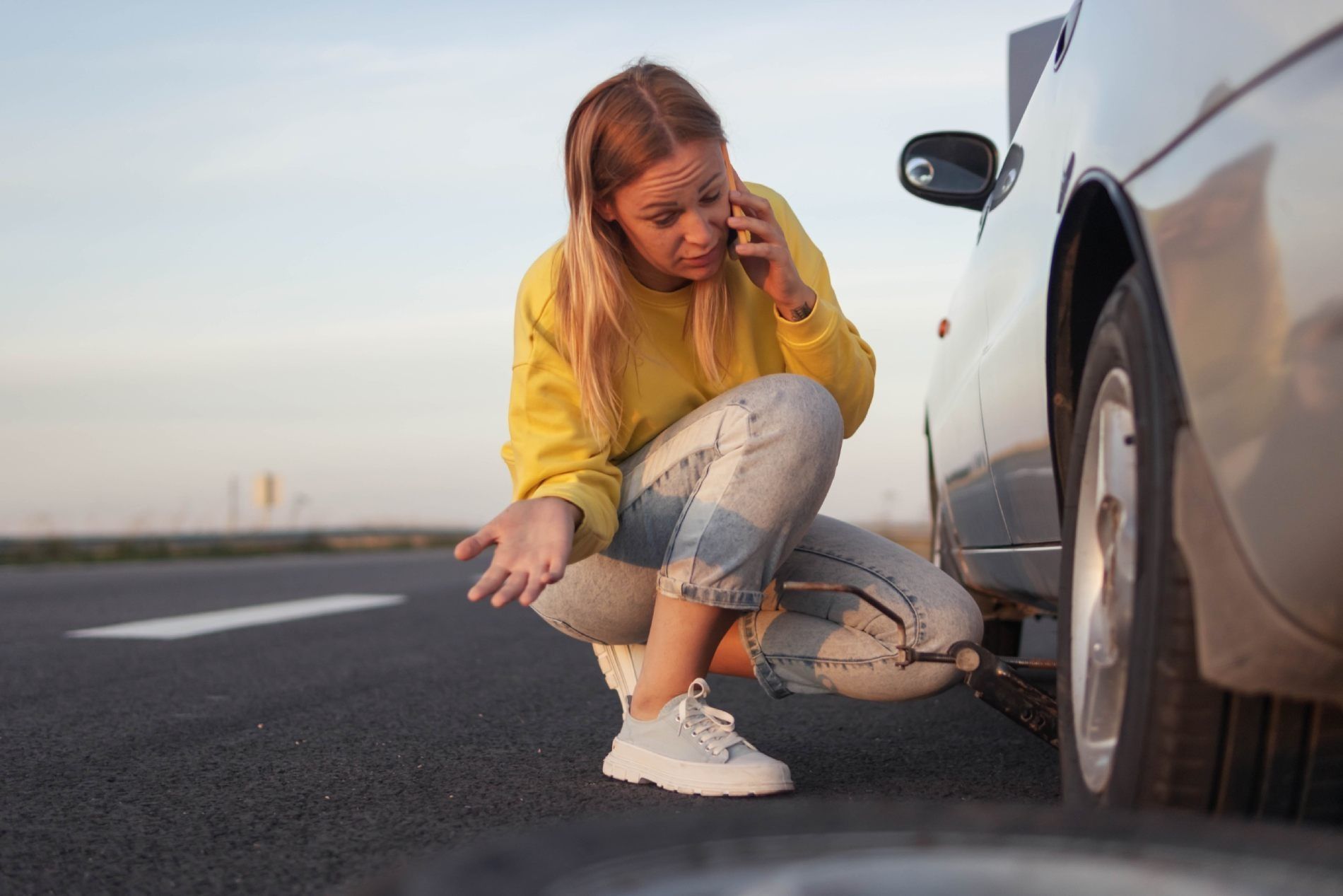Una mujer está arrodillada junto a un coche con una rueda pinchada y habla por un teléfono móvil.