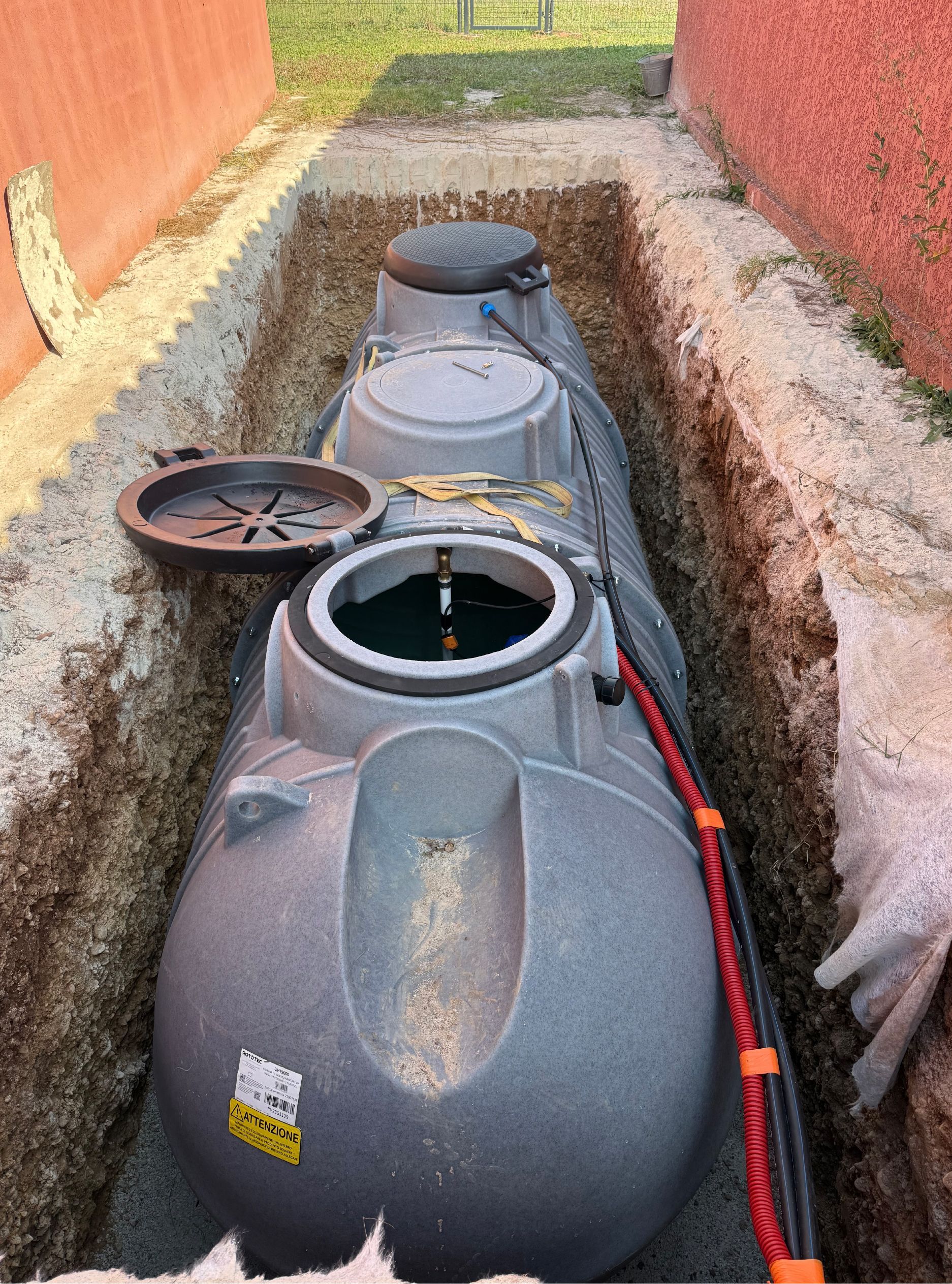 Cuve d'eau dans une tranchée étroite entre un mur rouge et le sol. Fosse grise avec couvercle ouvert.