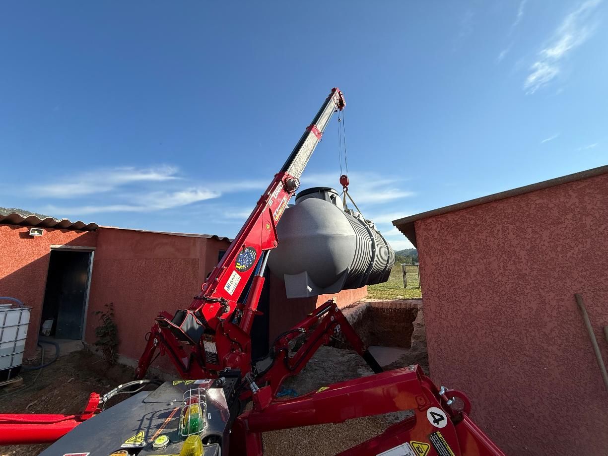 Grue rouge soulevant un grand réservoir gris près d'un bâtiment rouge sous un ciel bleu.