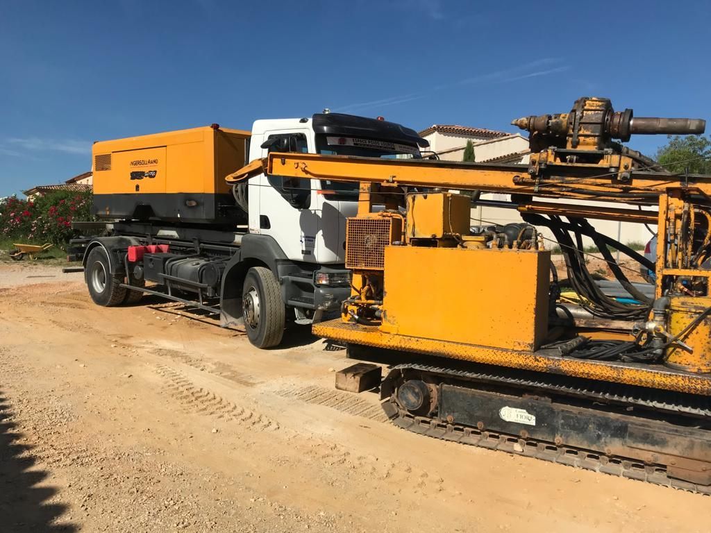 Appareil de forage jaune et blanc monté sur une plate-forme de camion garée sur un terrain en terre.