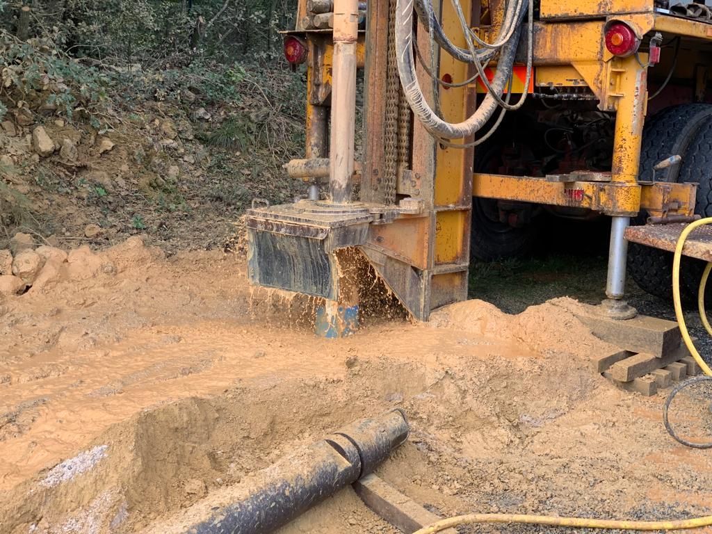 Foreuse creusant dans le sol, projetant de la terre. Décor extérieur avec un camion jaune.