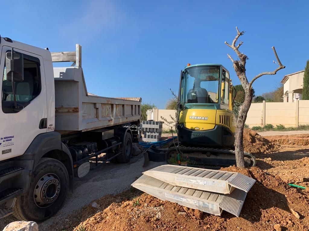 Scène de chantier avec un camion-benne, une pelleteuse jaune et des piles de blocs de béton. Journée ensoleillée.