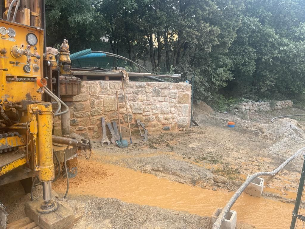 Foreuse jaune dans une zone boueuse près d'un mur de pierre avec outils. Arbres en arrière-plan.