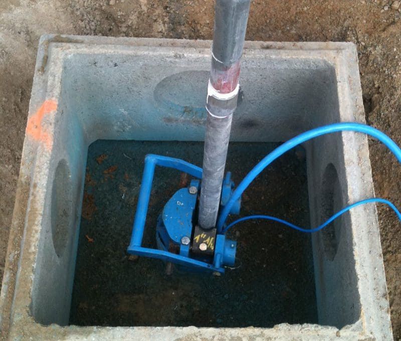 Pompe submersible bleue dans un puits en béton, reliée à des tuyaux et des fils.