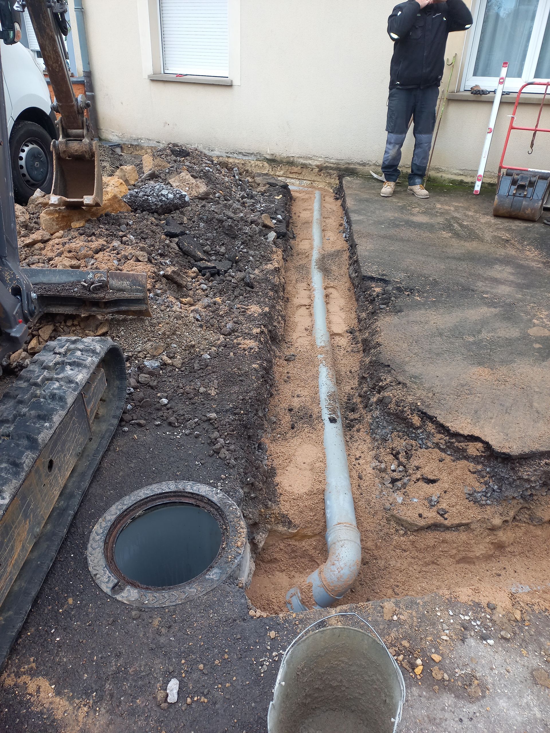 Chantier : tranchée avec tuyau gris, regard ouvert, ouvrier debout près d'une maison.
