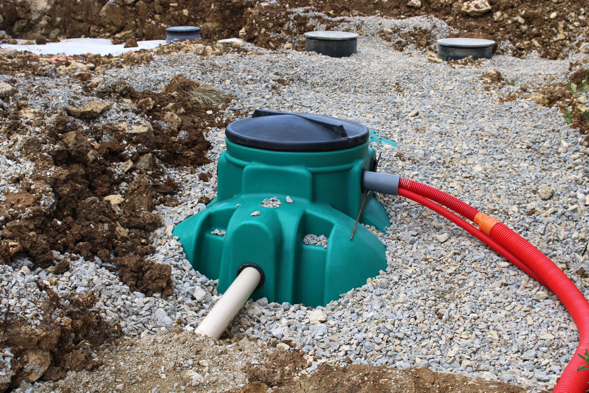 Fosse septique verte entourée de gravier, avec tuyaux rouges et blancs, et couvercle d'accès noirs.