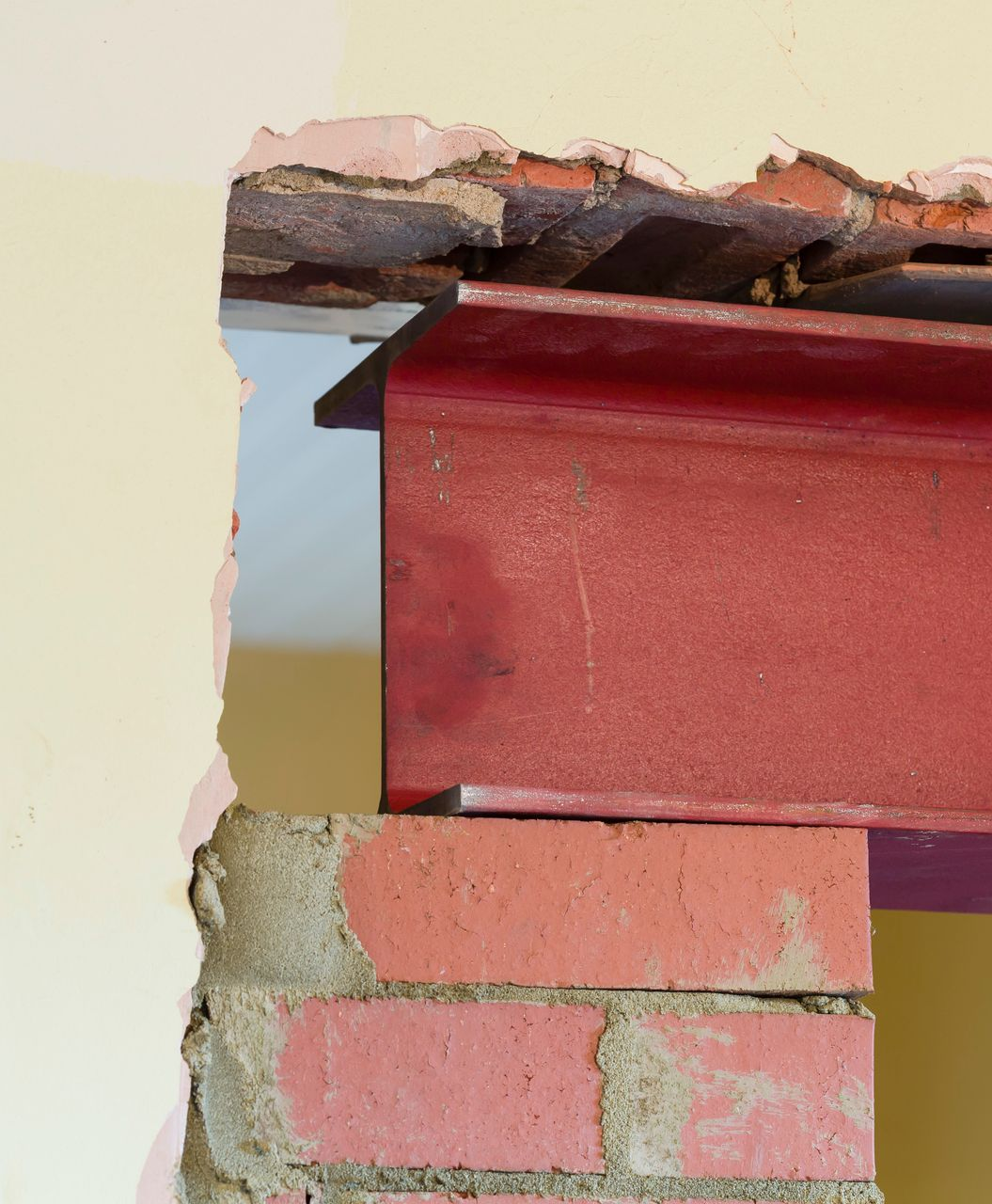 Poutre en acier rouge soutenant le plafond, posée sur des briques. Mur endommagé, signe de travaux.