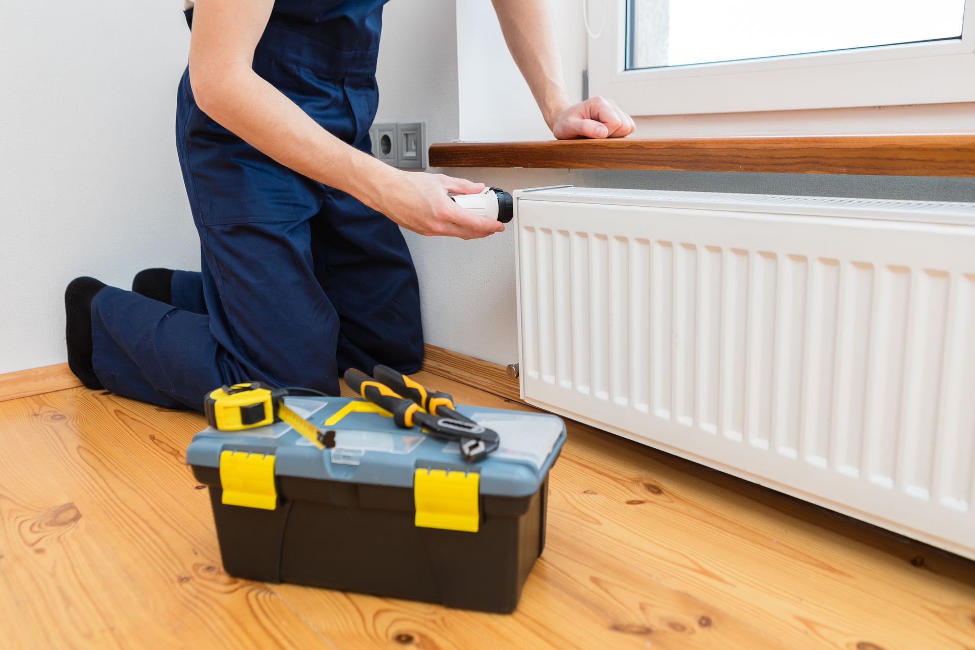 Personne agenouillée près d'un radiateur, travaillant dessus avec une boîte à outils et des outils à proximité.