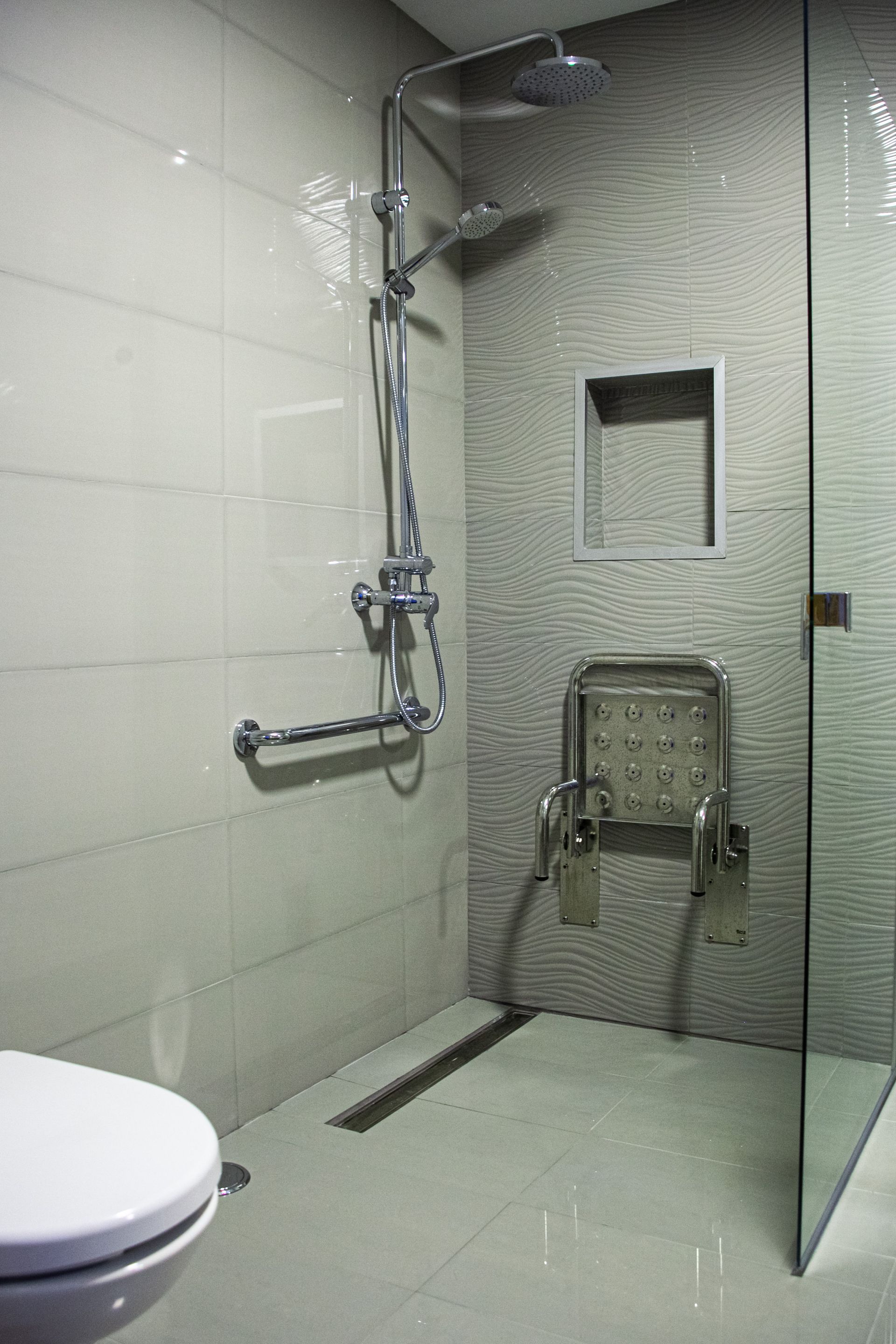 Salle de bains accessible : douche carrelée avec siège, barre d'appui et pommeau de douche à main, porte vitrée.