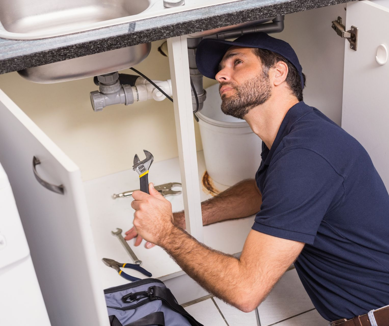 Plombier travaillant sous un évier de cuisine, tenant une clé, regardant vers le haut.