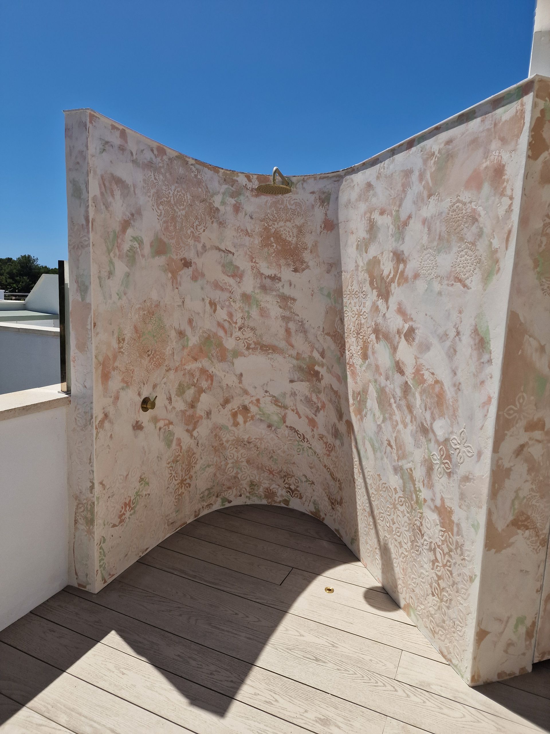 Ducha al aire libre con paredes curvas, piso de madera y cabezal de ducha de latón contra un cielo azul.