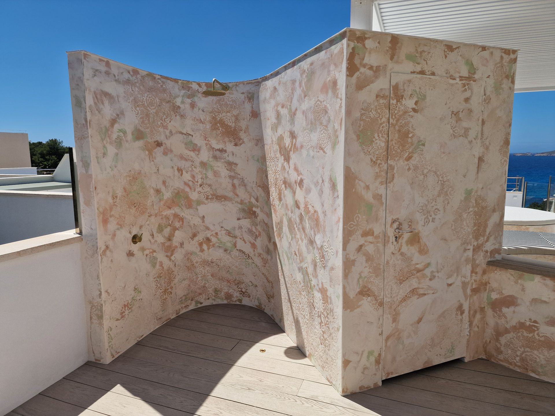 Mampara de ducha exterior con paredes texturizadas en color tostado y suelo gris, con vistas al océano.