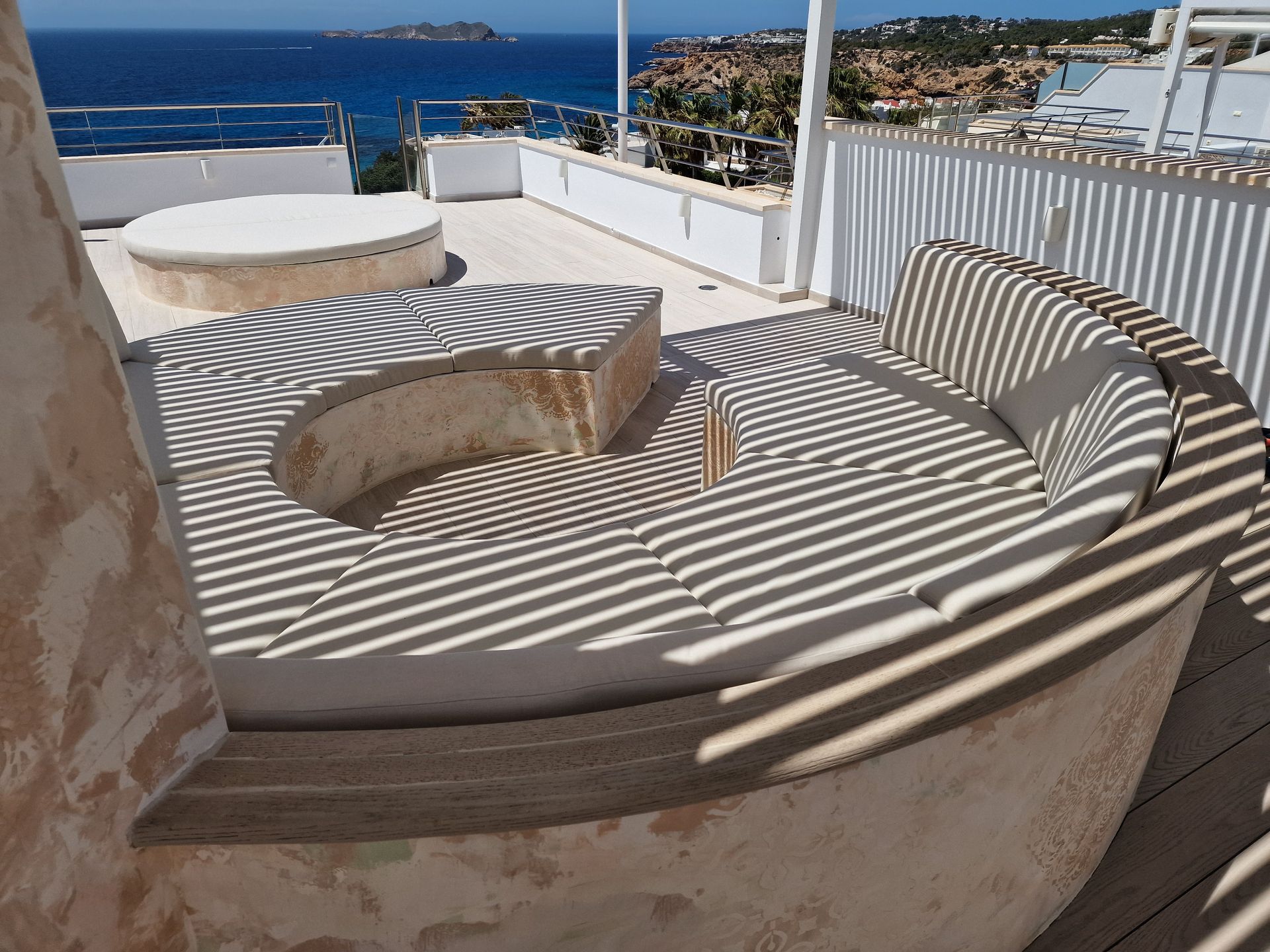 Zona de estar al aire libre con vista al mar; banco de concreto curvo con cojines, terraza, barandilla blanca.