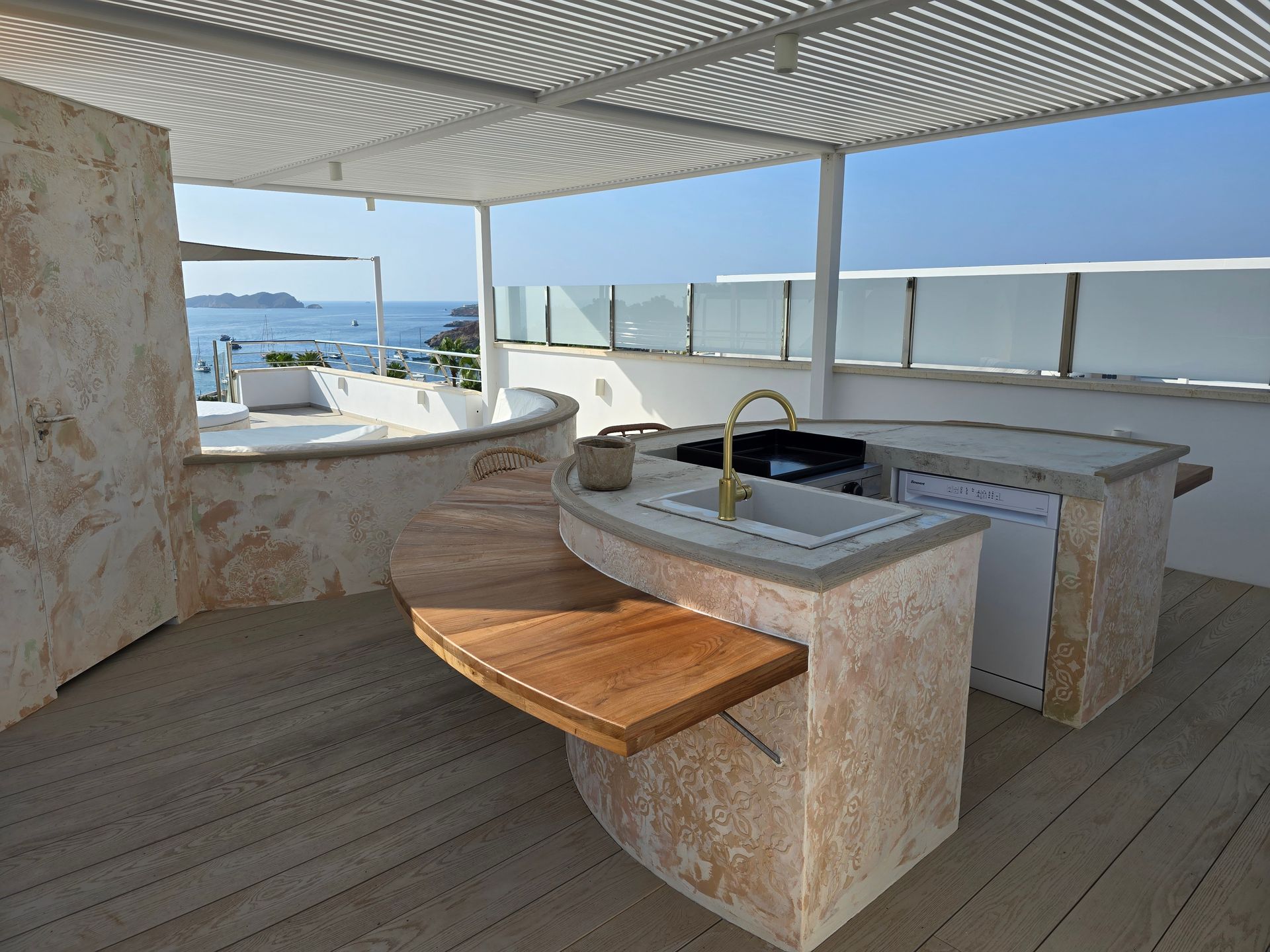 Cocina al aire libre con encimeras de piedra, fregadero, parrilla y tabla de cortar de madera, con vista al mar.