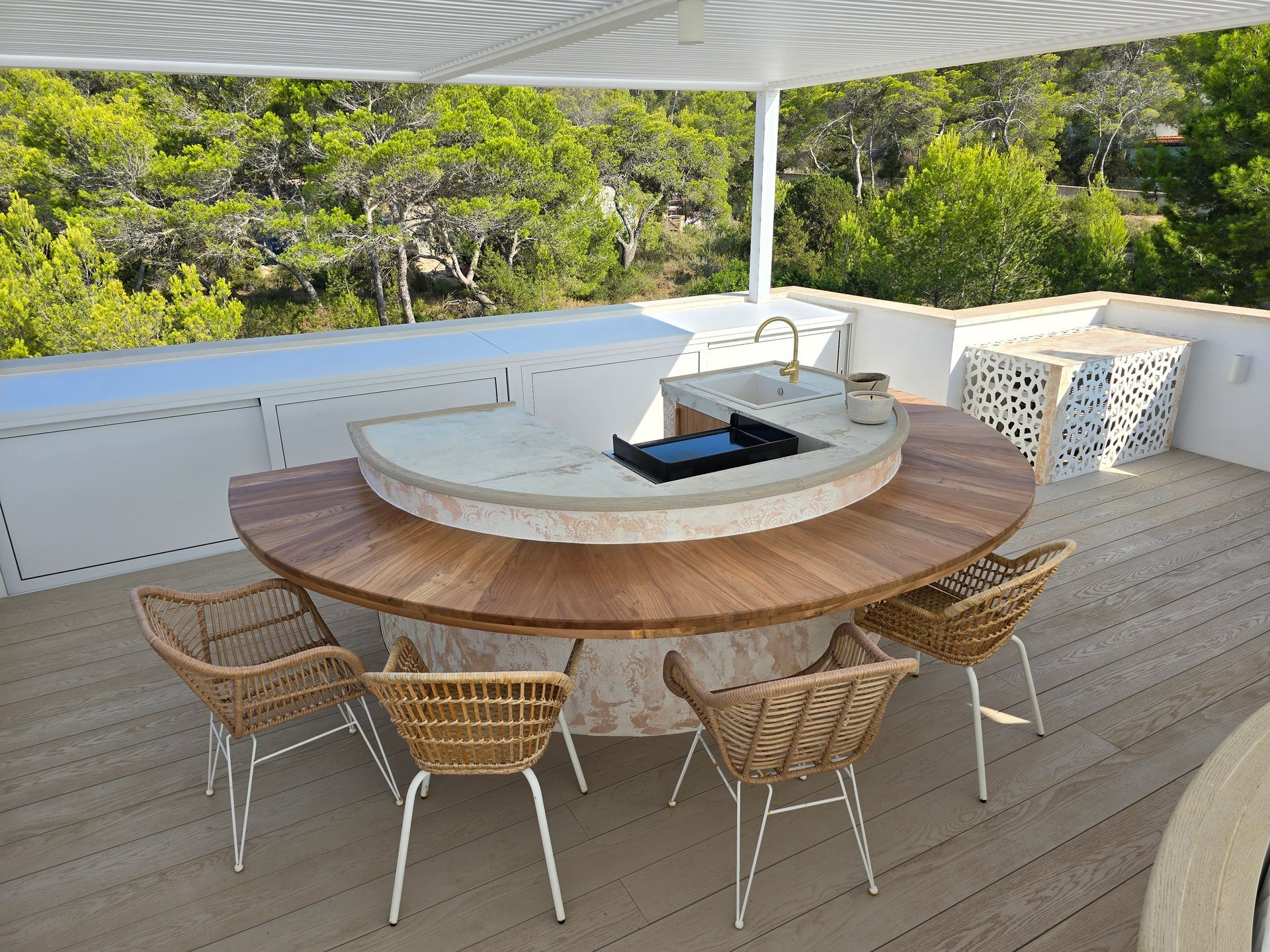 Cocina al aire libre con mesa de madera, encimera circular con parrilla, sillas de mimbre y fondo de bosque.