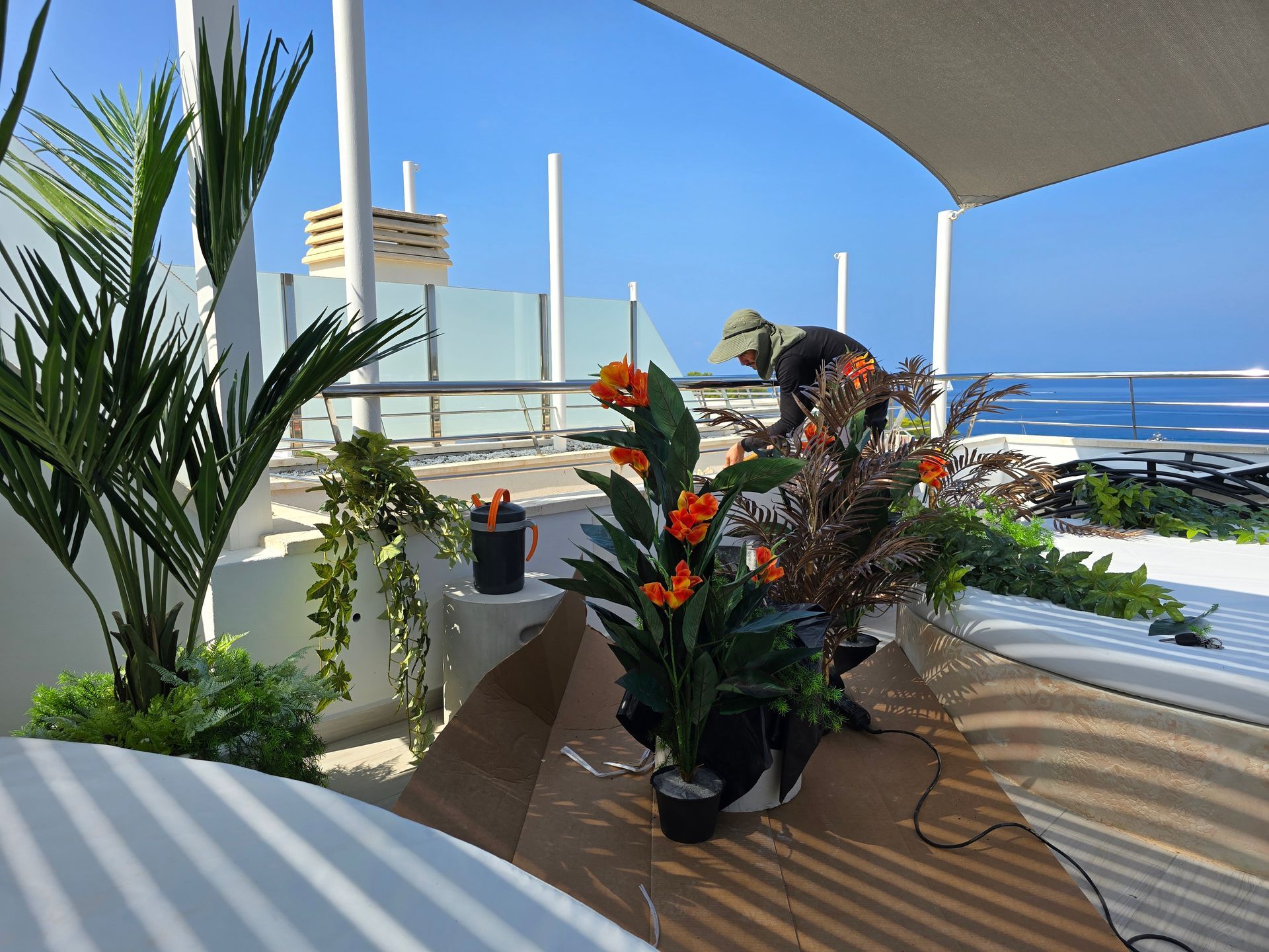 Persona cultivando un huerto en un patio con plantas y vista al mar. Día soleado, muebles blancos y sombrilla color canela.