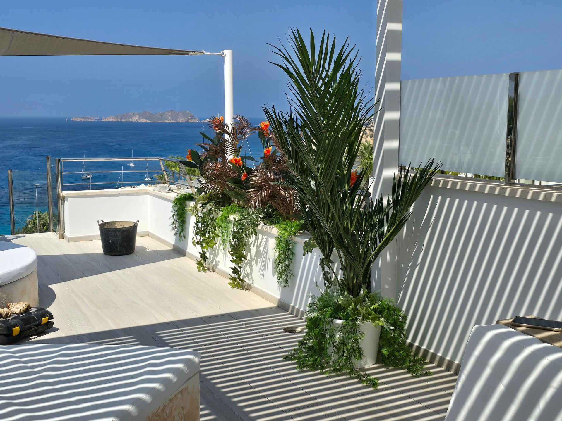 Terraza con vistas al mar, con plantas, estructura de sombra y agua azul brillante de fondo.
