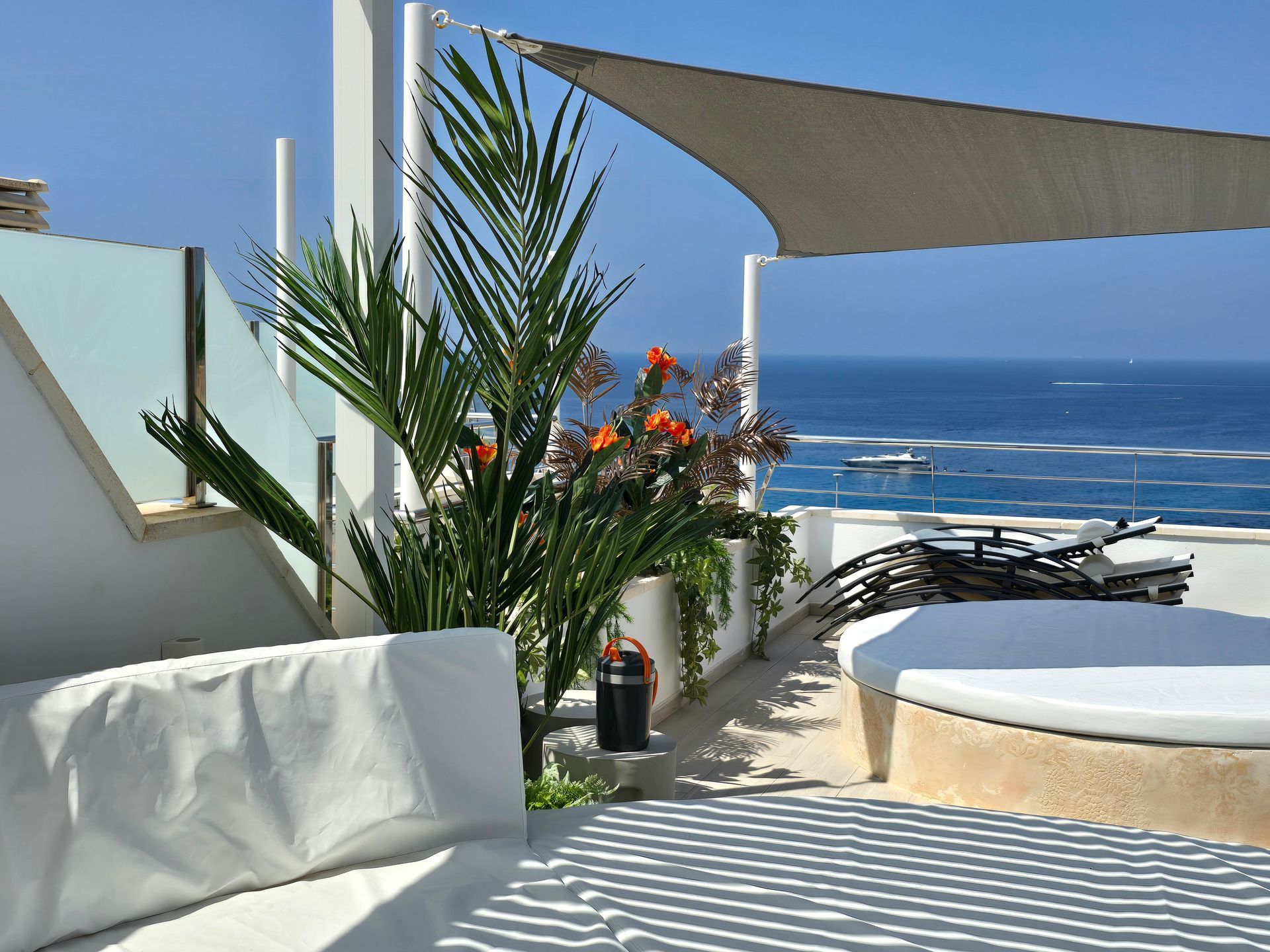 Patio en la azotea con vista al mar, cojines blancos, palmeras, velas de sombra, agua azul, día soleado.