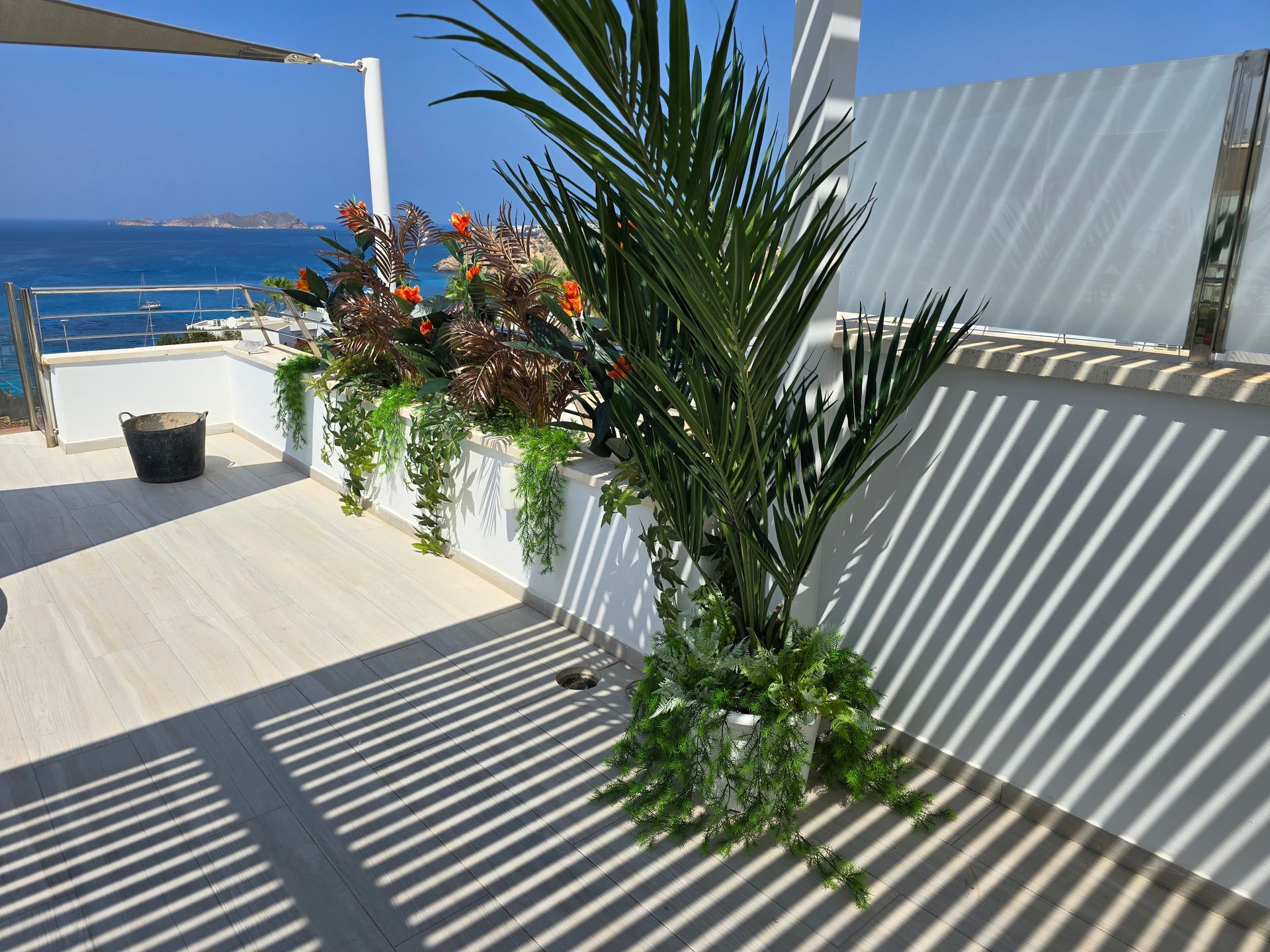 Terraza con vista al mar, palmeras y plantas colgantes, proyectando sombras.