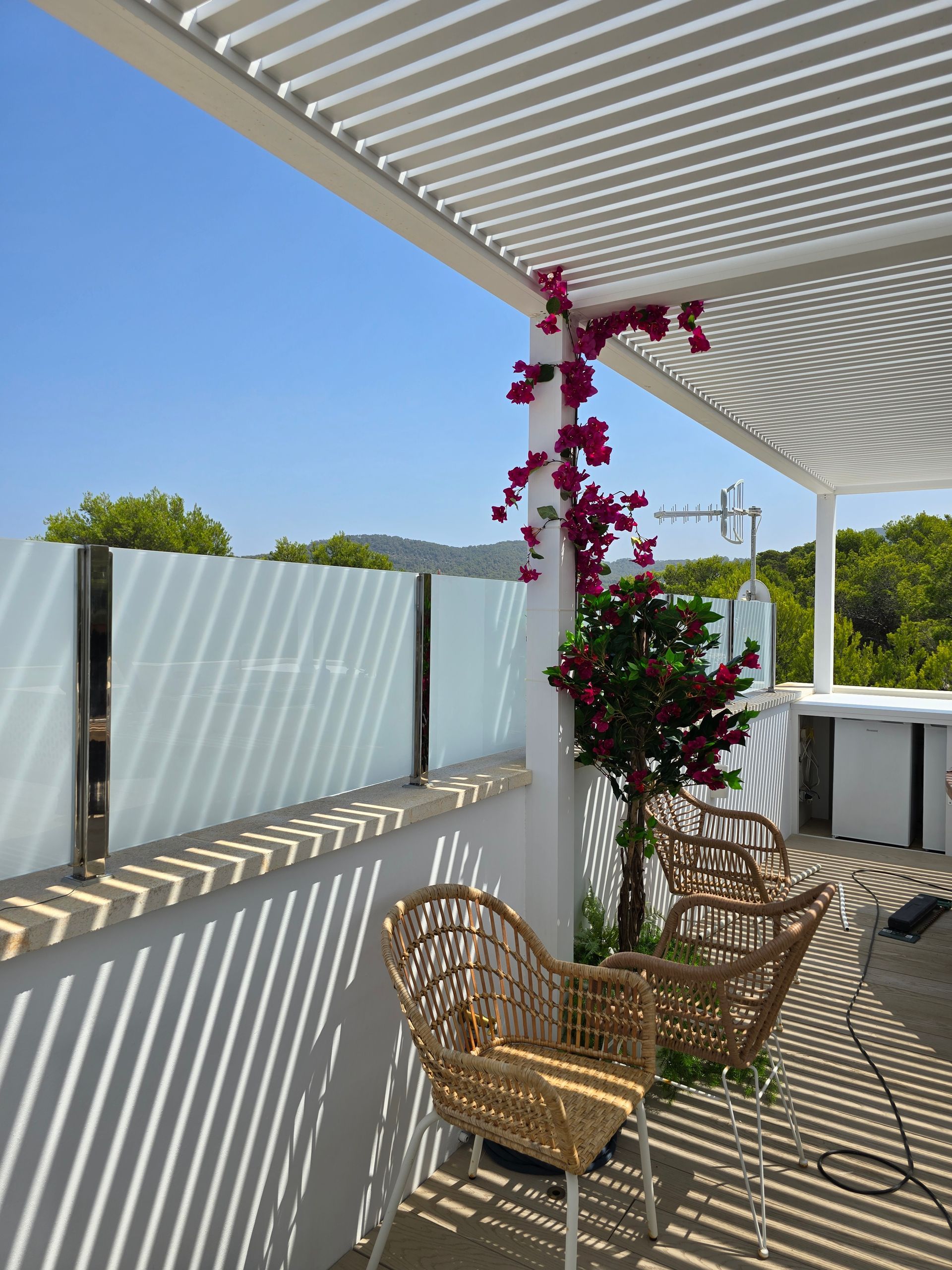 Patio exterior con dos sillas de mimbre, pérgola y buganvillas bajo un cielo azul.