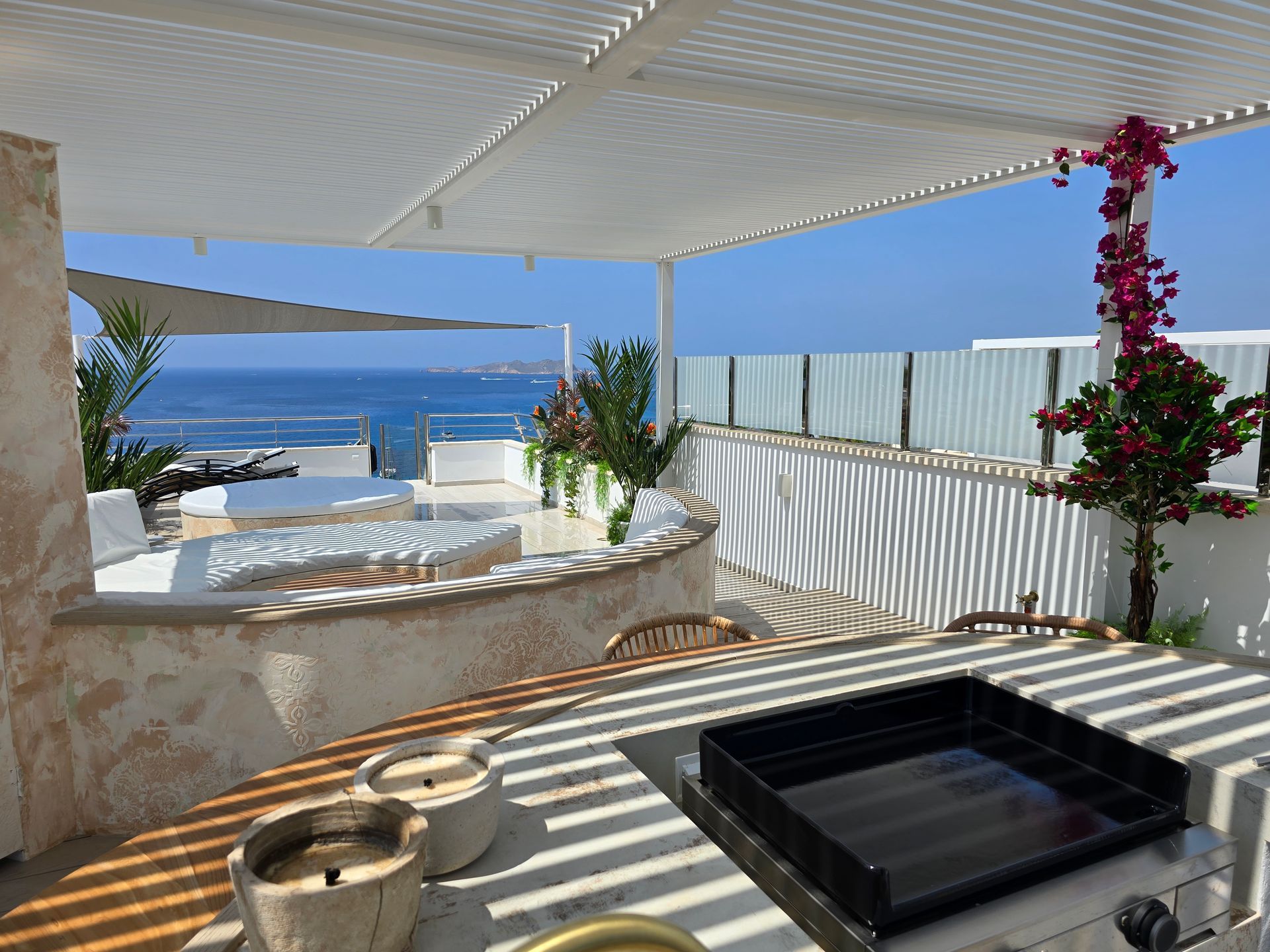 Patio al aire libre con vista al mar, pérgola blanca, parrilla, asientos incorporados y buganvillas.