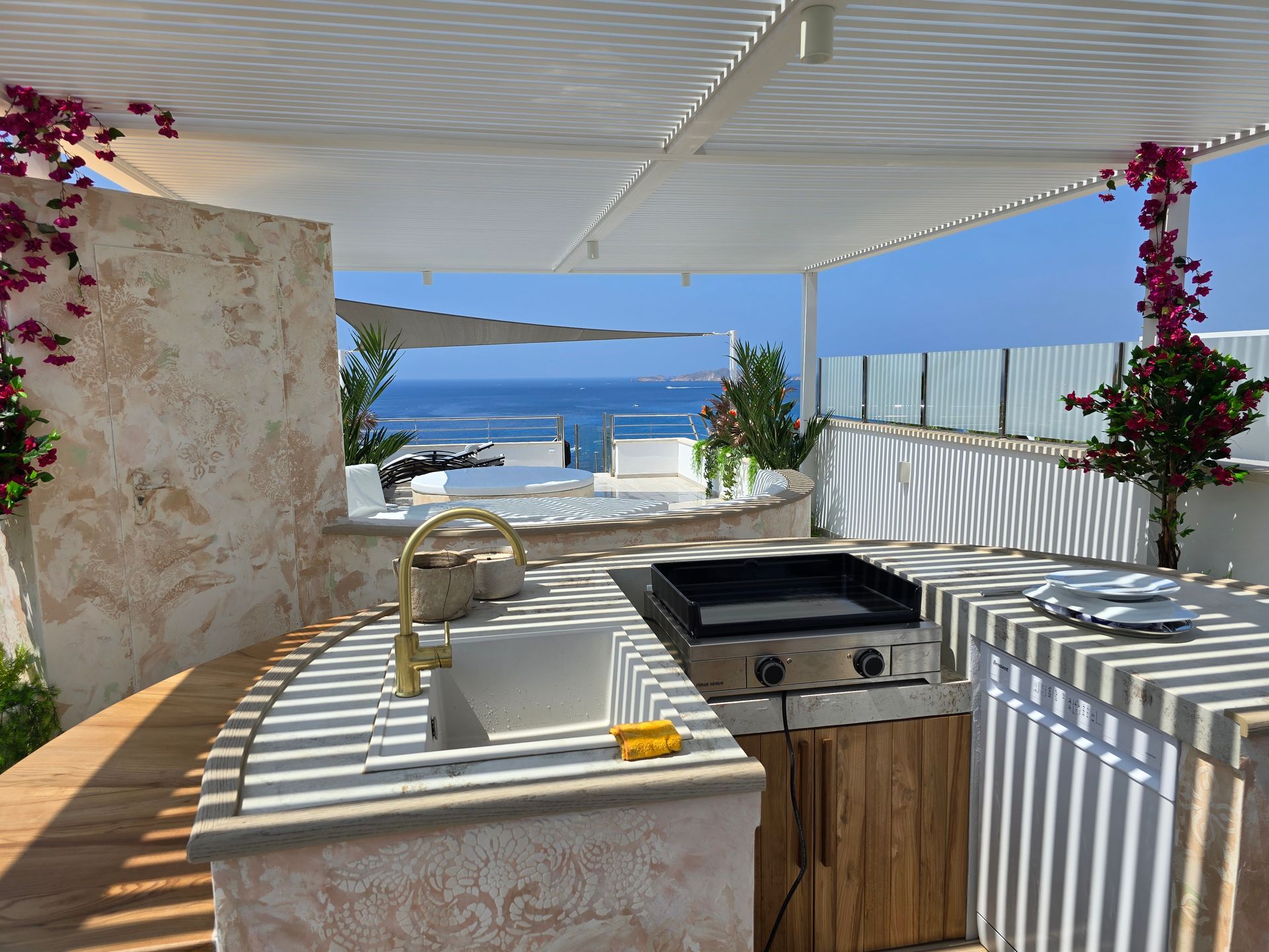Cocina exterior con vista al mar, parrilla, fregadero y buganvillas en un día soleado.