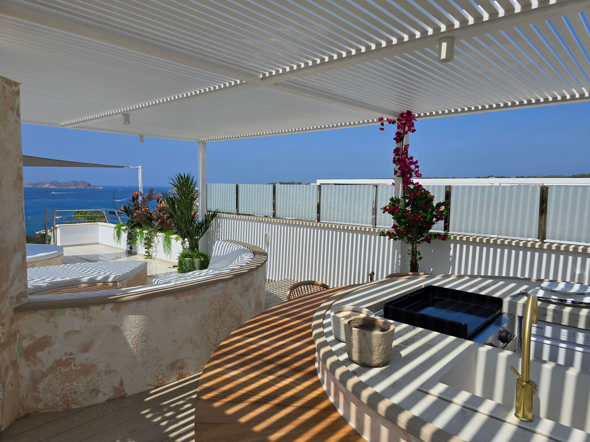 Patio exterior con pérgola blanca, vista al mar, parrilla incorporada y plantas decorativas.
