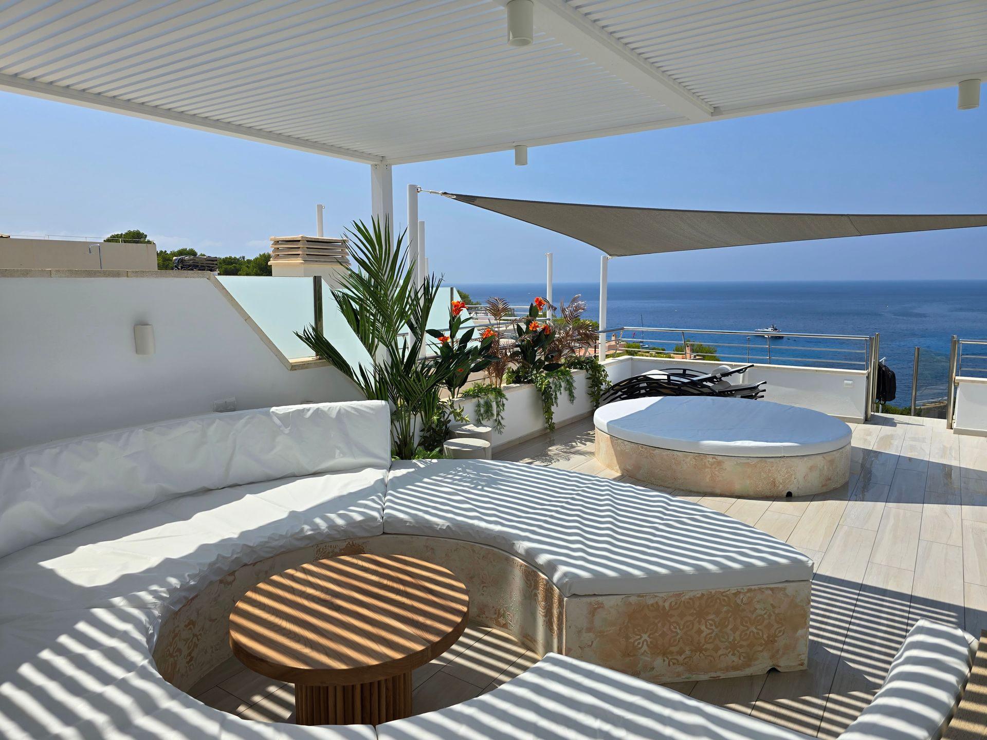 Terraza en la azotea con vistas al mar. Asientos blancos, mesa redonda, jacuzzi y zona de sombra. Día soleado.