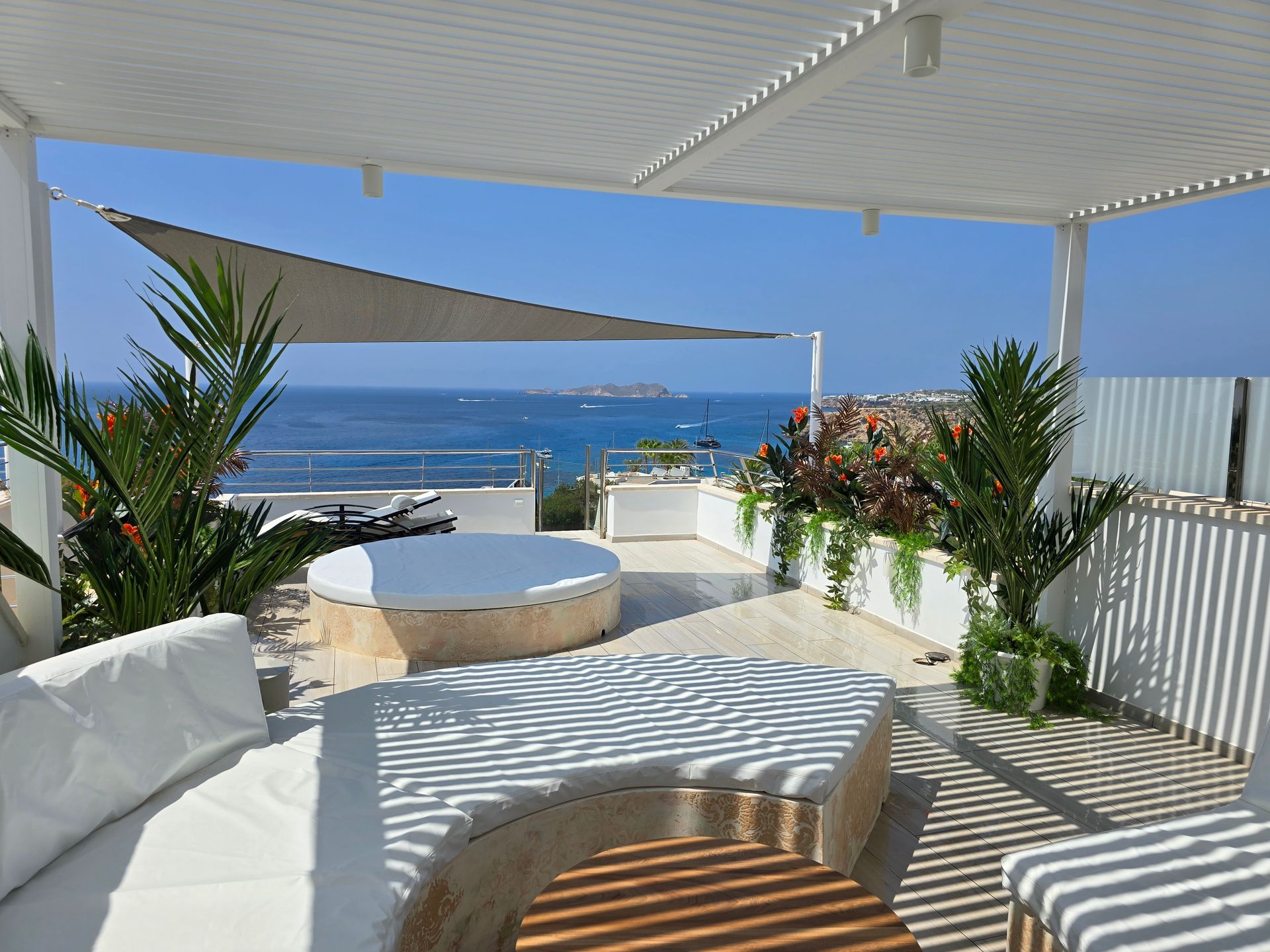 Patio en la azotea con muebles blancos, vista al mar, cielo azul y plantas tropicales.