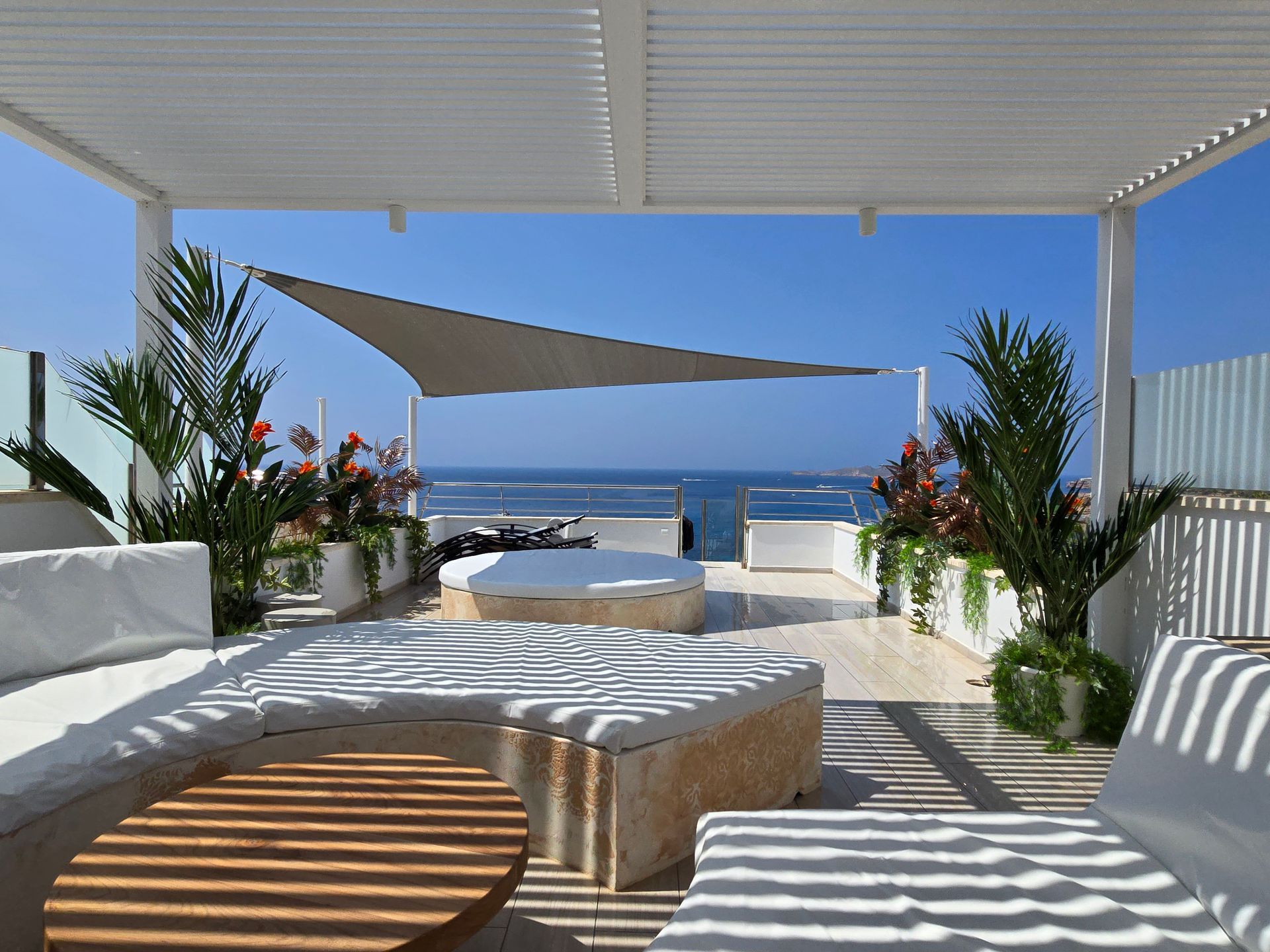 Patio en la azotea con muebles blancos, mesas de madera, toldo y vista al mar.