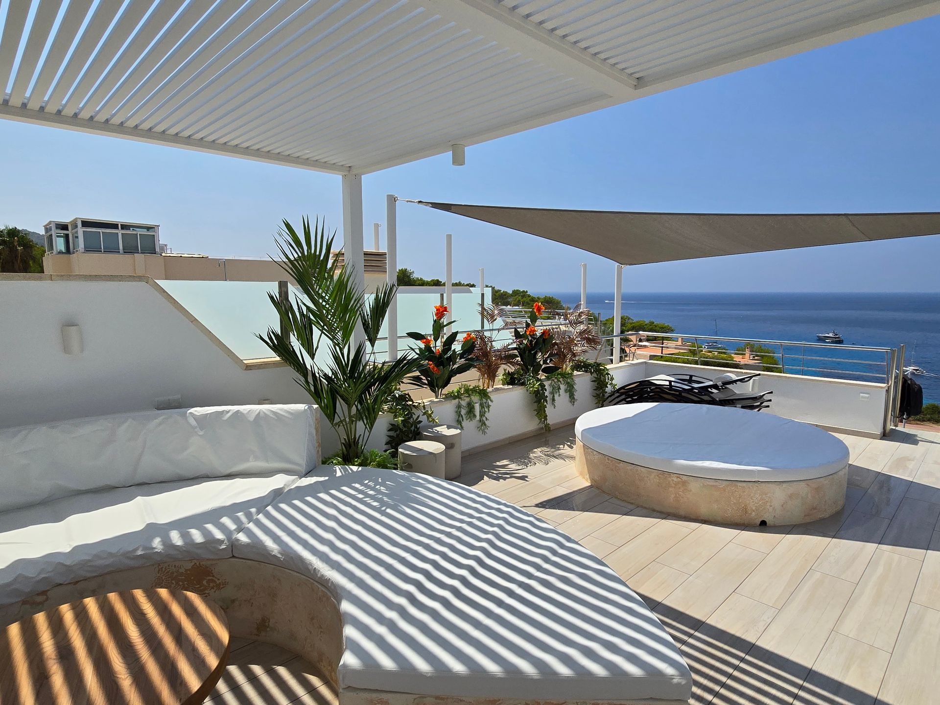 Patio en la azotea con vistas al mar, tumbonas, velas de sombra y pérgola. Cielo azul y agua.
