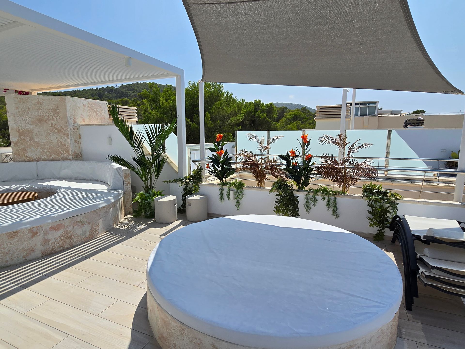 Patio en la azotea con un diván redondo cubierto de blanco, un pozo de fuego y plantas en macetas bajo una vela de sombra.