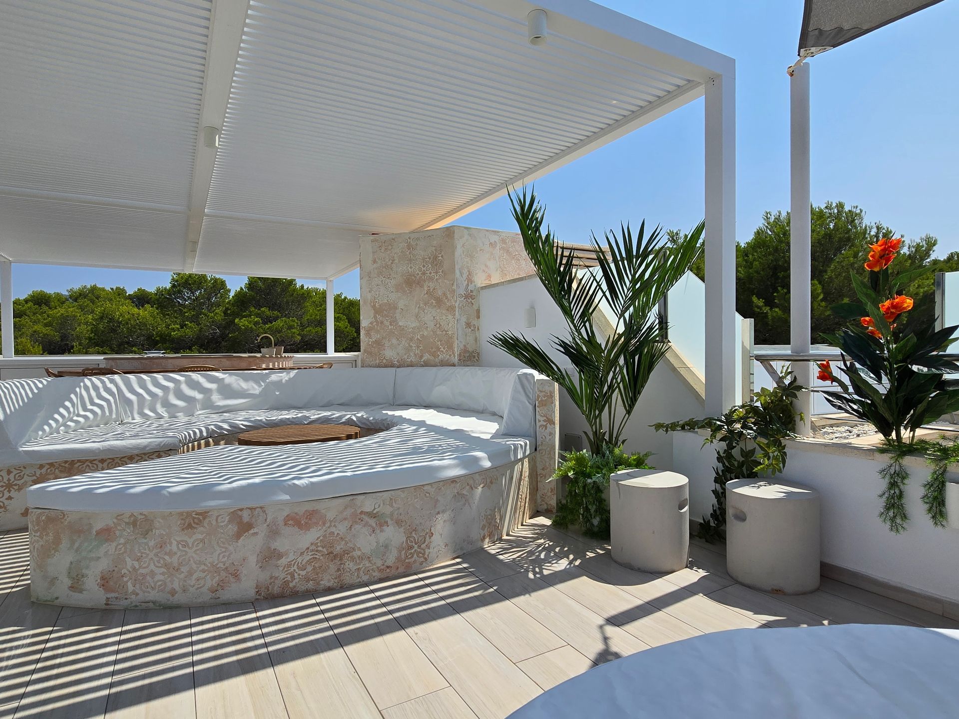 Patio en la azotea con pérgola blanca, asientos empotrados, plantas y fogata. Día soleado.