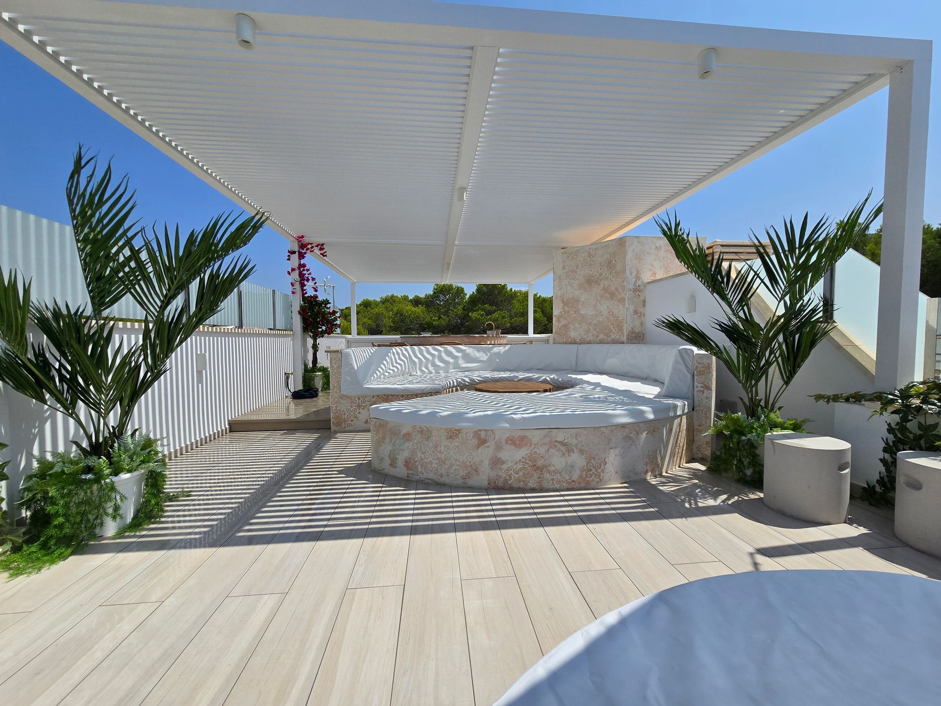 Patio en la azotea con pérgola blanca, asientos de piedra, plantas y una terraza de madera clara bajo un cielo azul.