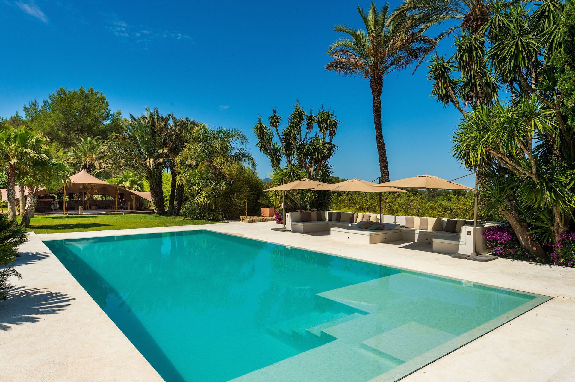 Piscina rectangular de agua turquesa, patio con sombrillas y exuberante vegetación bajo un cielo azul.