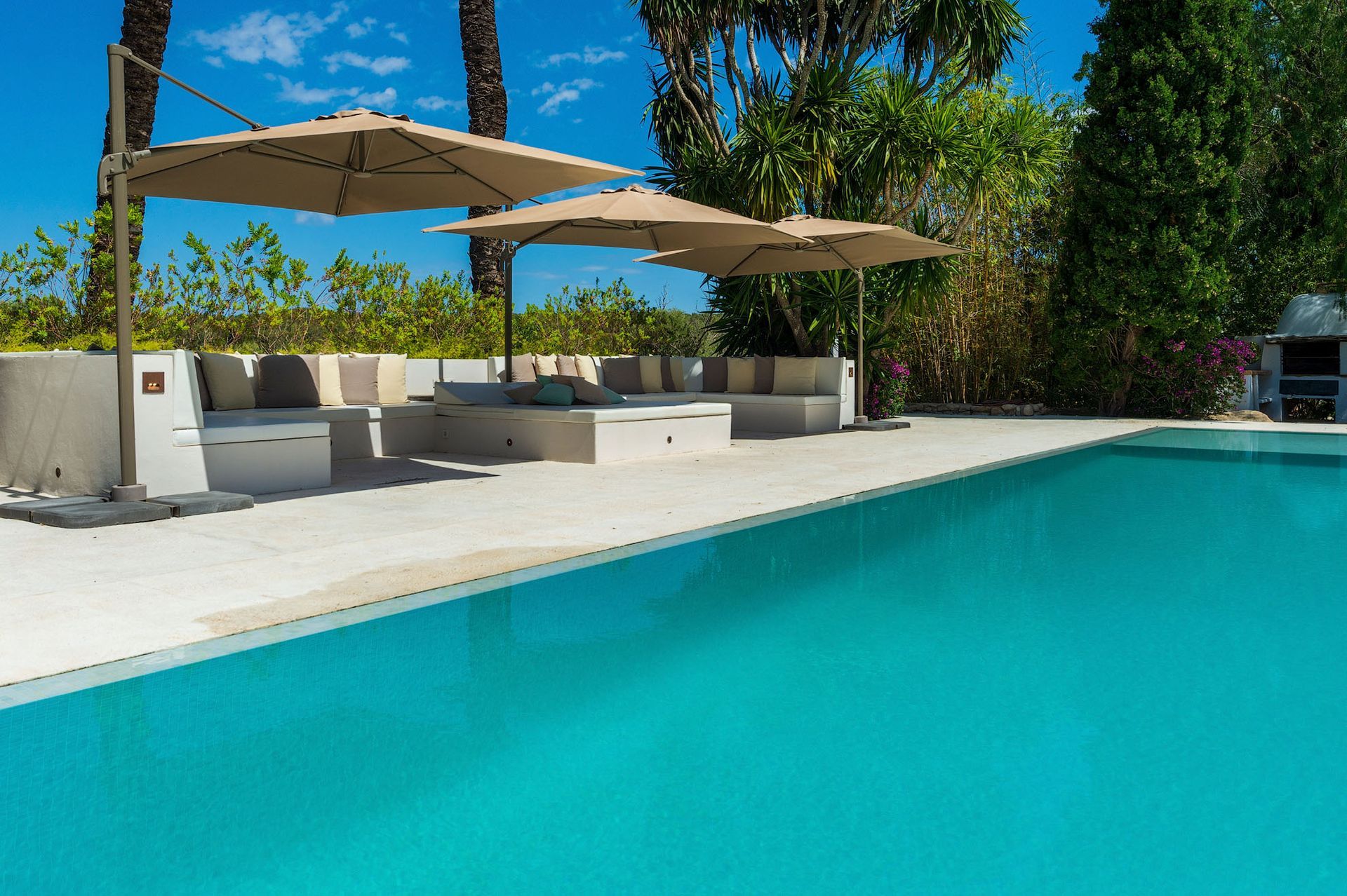 Zona de salón junto a la piscina con sombrillas, asientos de colores claros y agua turquesa bajo un cielo azul.