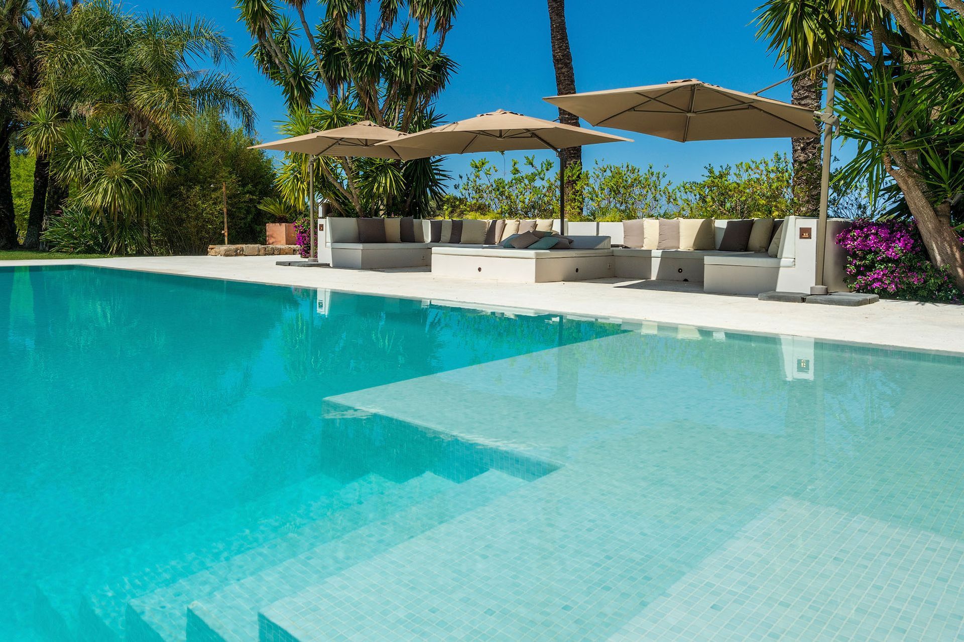 Piscina con agua azul claro y escalones, tumbonas blancas con sombrillas y exuberante follaje verde.