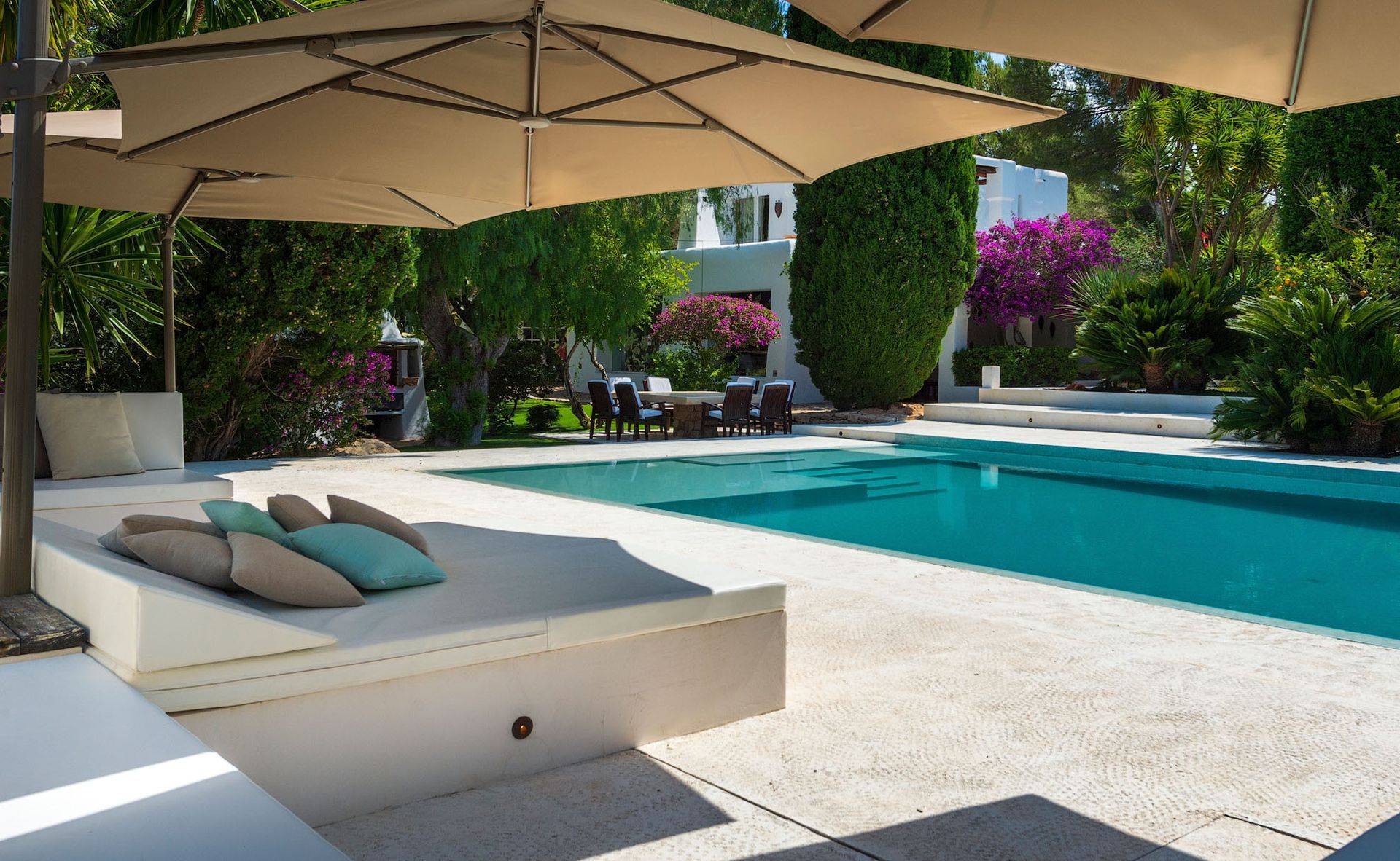 Área de descanso junto a la piscina con sombrillas, almohadas y vista a la piscina y los árboles.