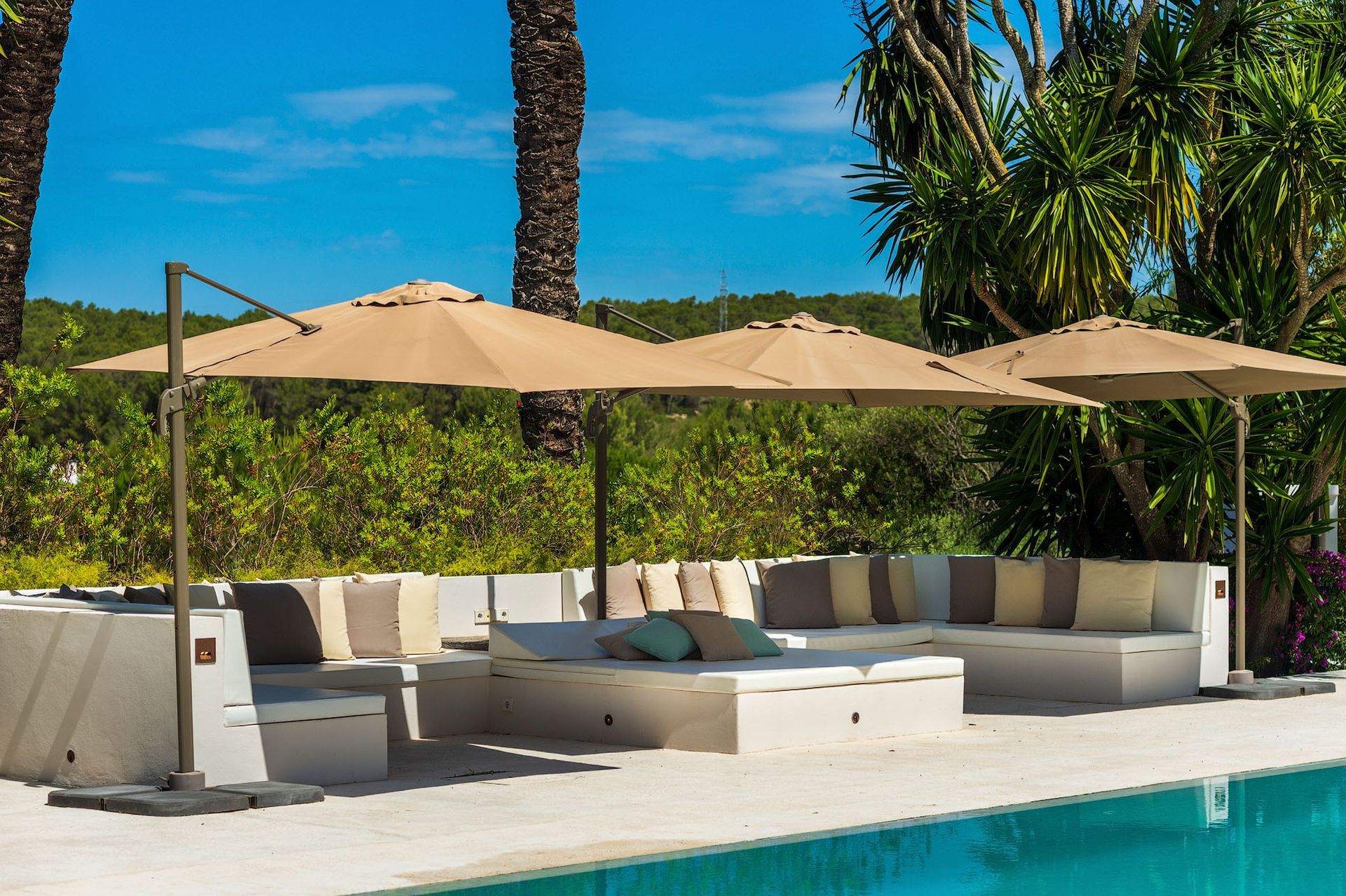 Zona de salón junto a la piscina con sombrillas beige que dan sombra a los sofás blancos con cojines.