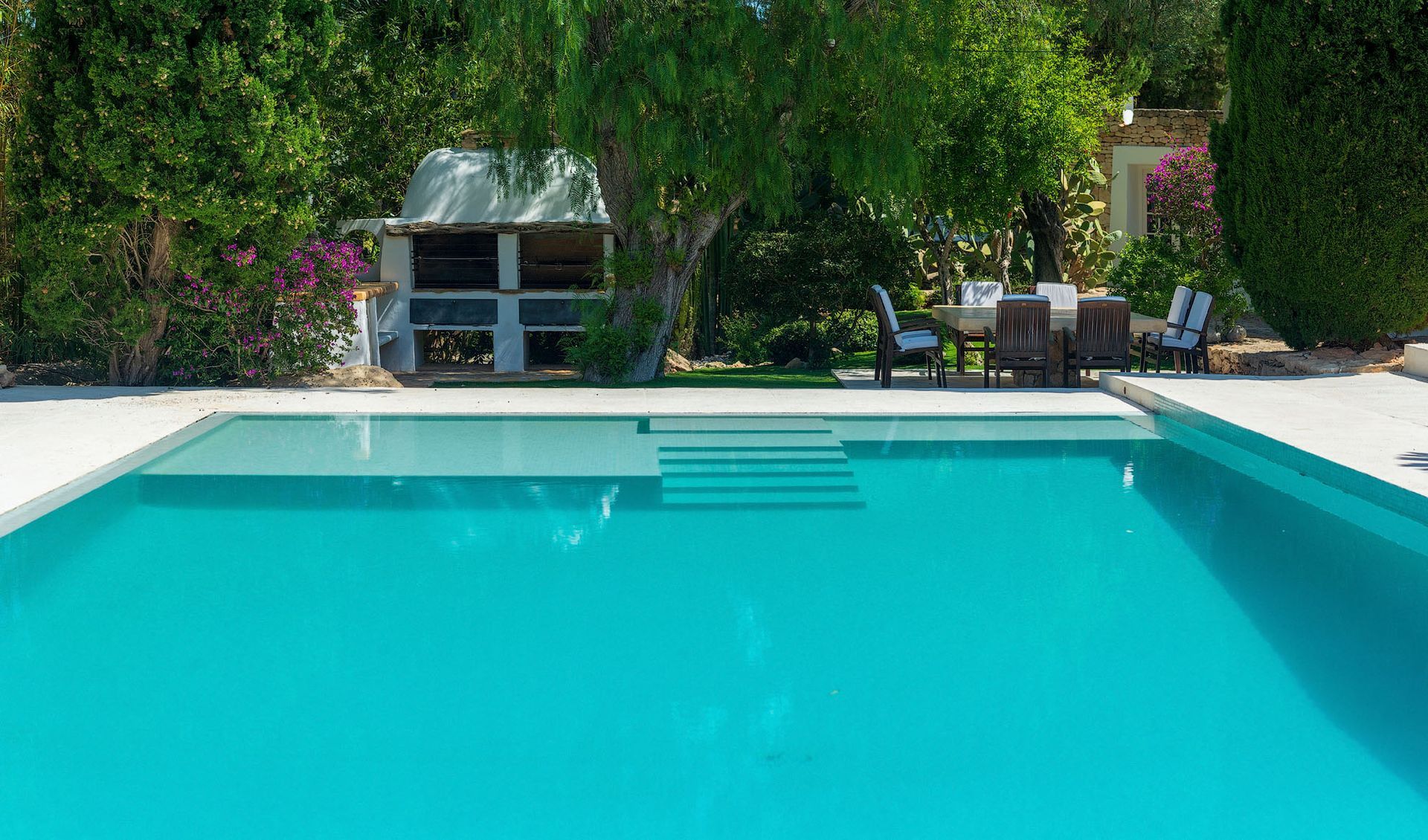 Piscina con agua turquesa, escaleras y zona de patio circundante con árboles y muebles de exterior.