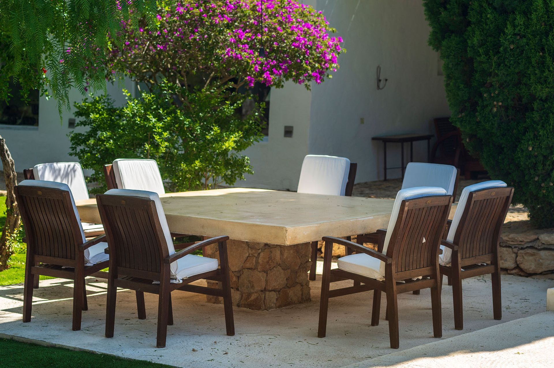 Comedor de exterior bajo un árbol de flores moradas. Mesa cuadrada de piedra con ocho sillas de madera y cojines blancos.