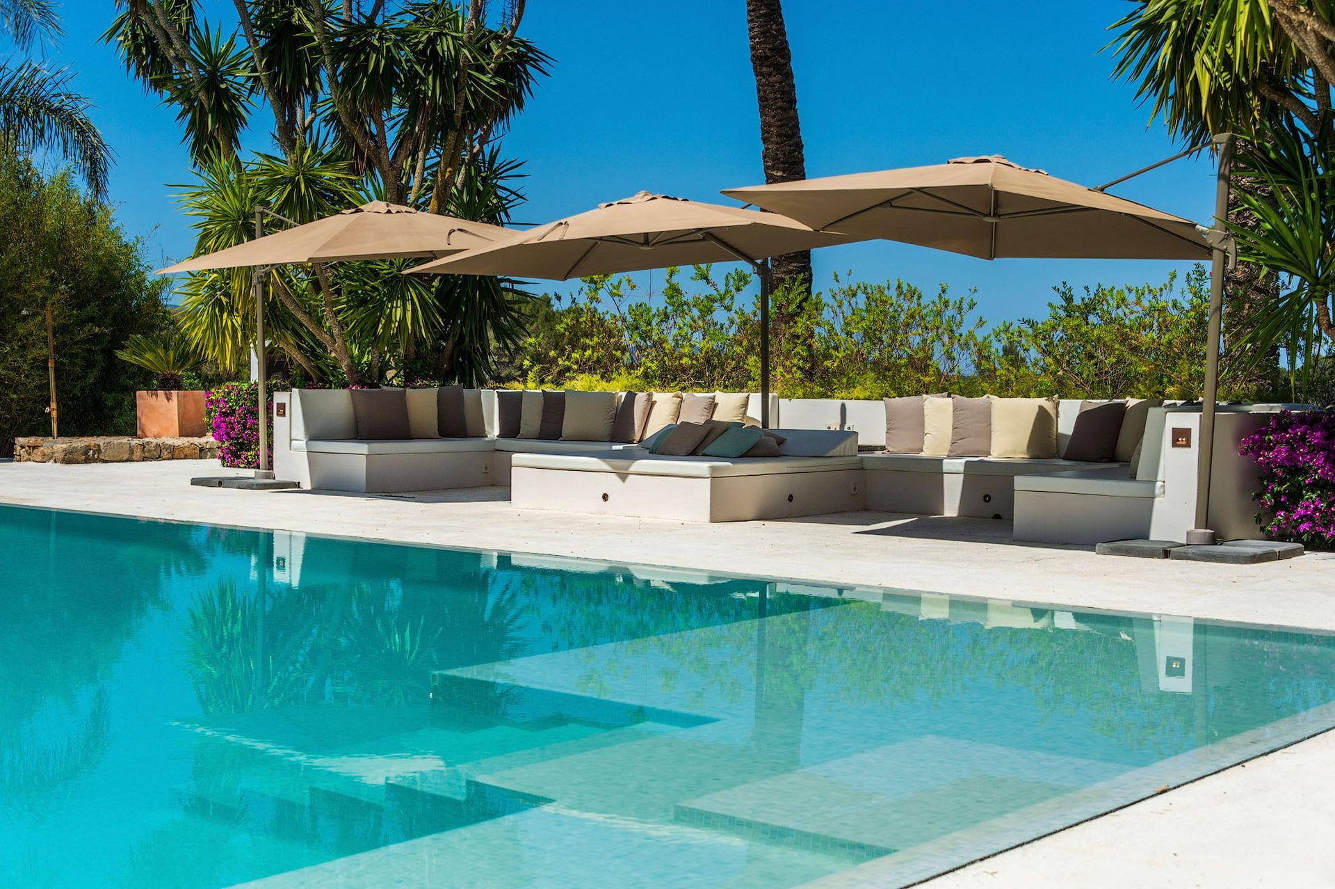 Área de descanso junto a la piscina con sombrillas, asientos de colores claros y agua azul cristalina.
