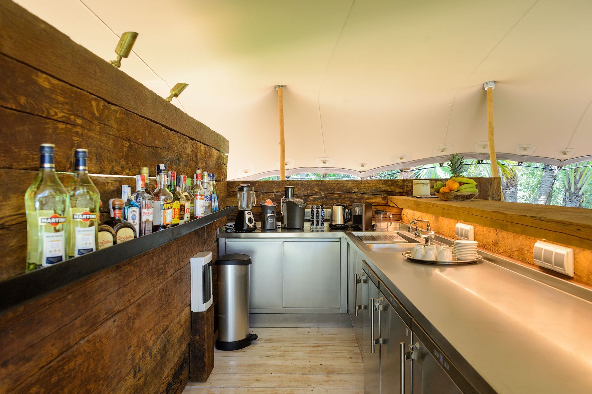 Zona de bar dentro de una carpa. Barra de madera con botellas, mostrador de acero inoxidable con electrodomésticos.